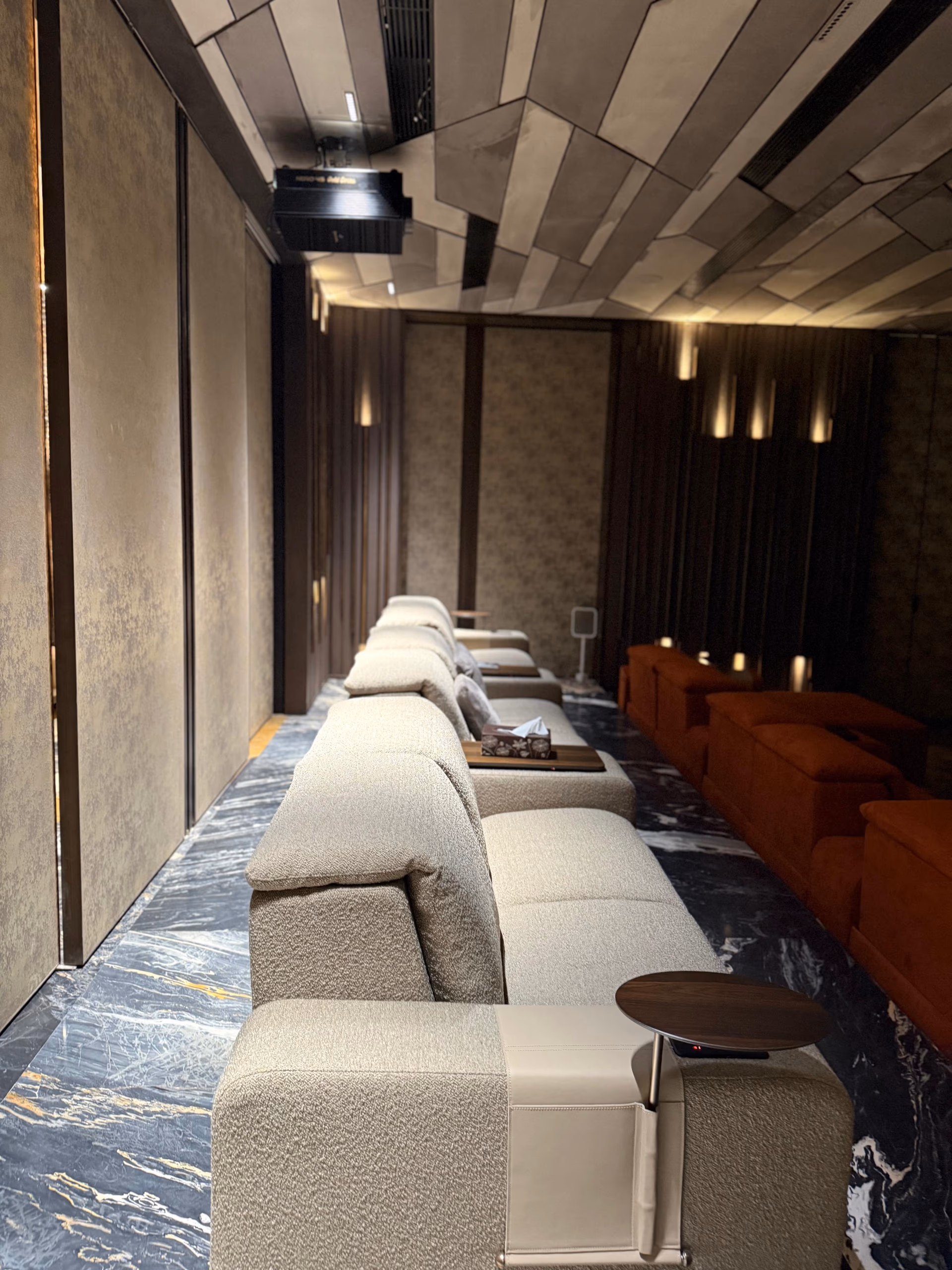 Row of cream boucle theater recliners with swing-out side tables, facing a second orange sofa row inside a dark panelled home theater.