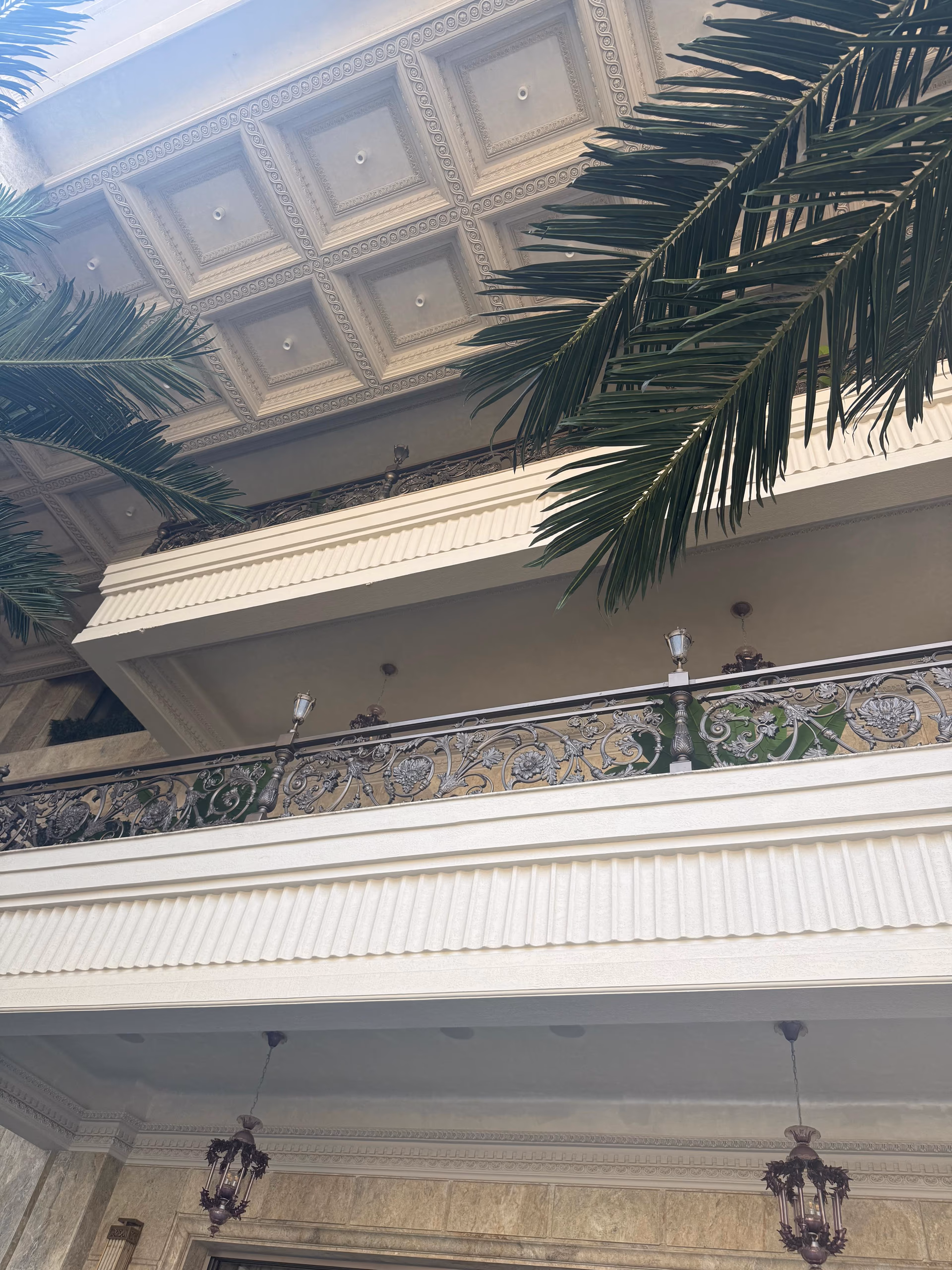 Upward view of stacked balconies with carved cornices, wrought-iron floral railings, and lantern pendant lights against a palm frond.