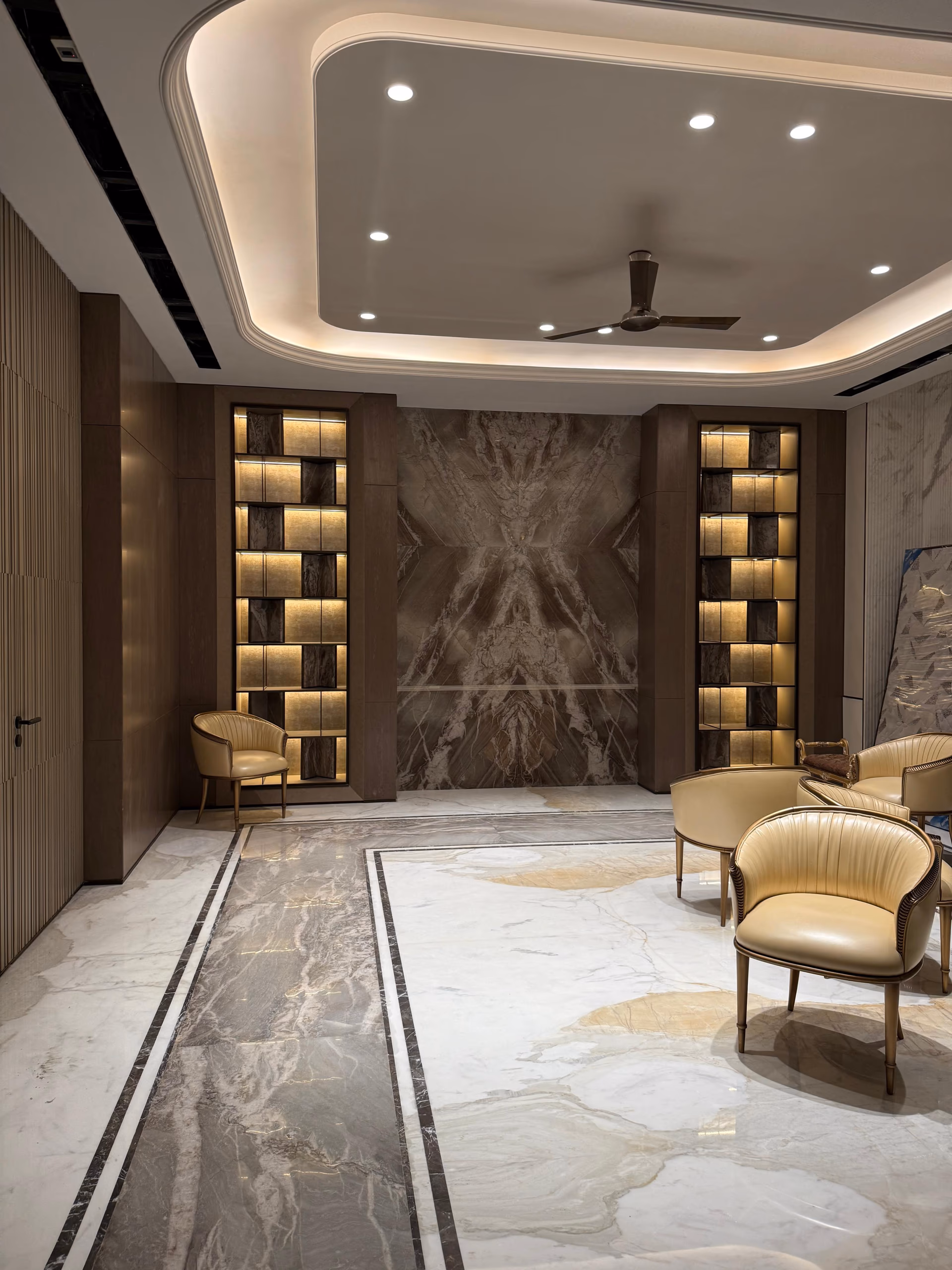 Lounge seating area with gold leather barrel chairs, a bookmatched marble feature wall, and twin backlit display shelving columns.