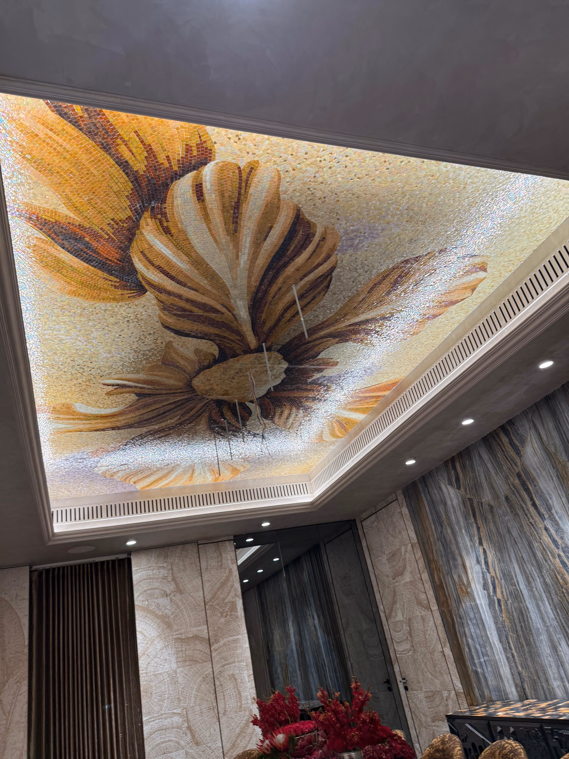 Backlit glass mosaic ceiling artwork of a golden feather or shell motif above a mirrored corridor with fluted marble panelling.