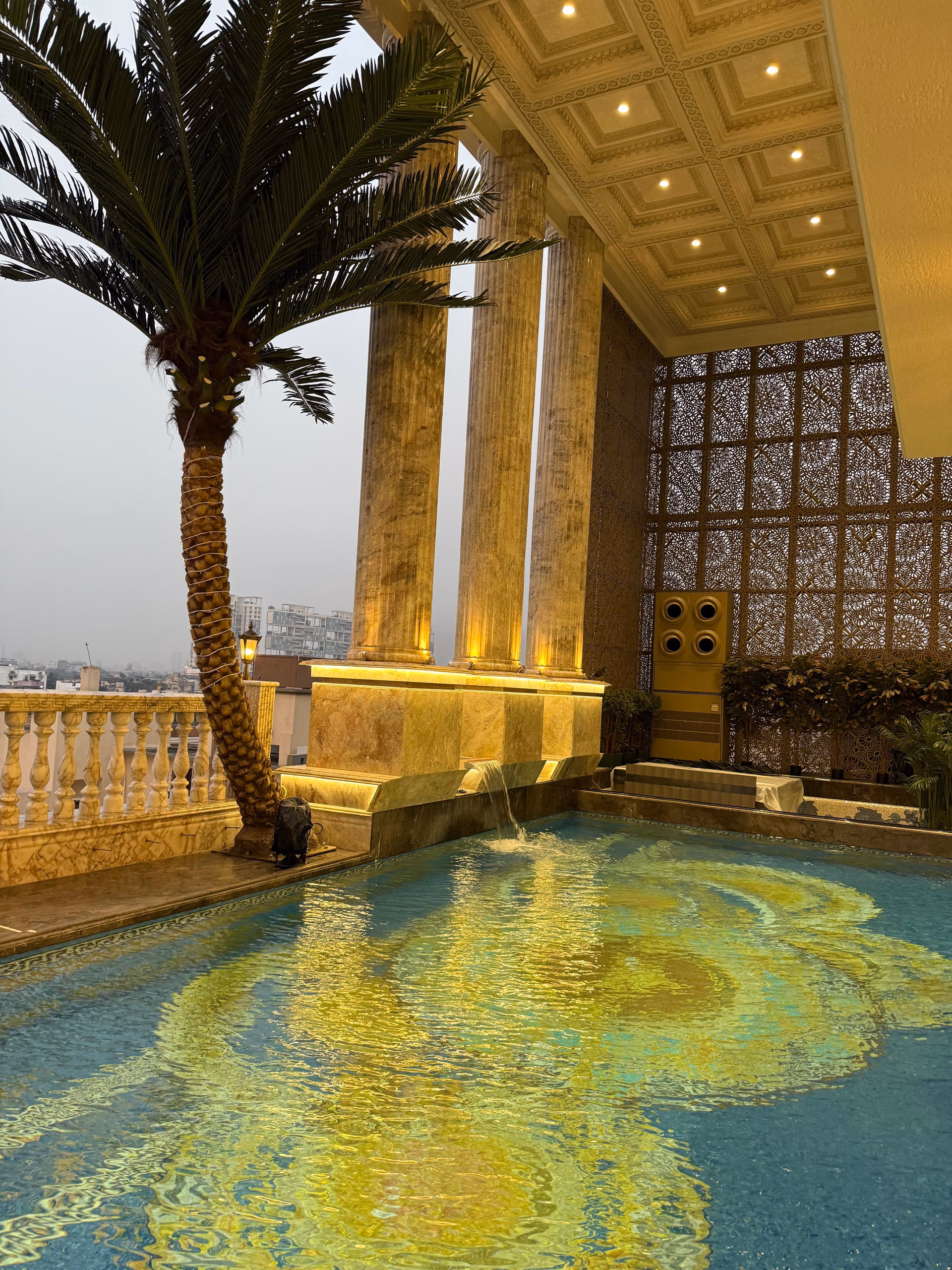 Dusk view of a rooftop terrace with tall fluted columns, a coffered gold ceiling, a jali screen wall, and a lit pool below.