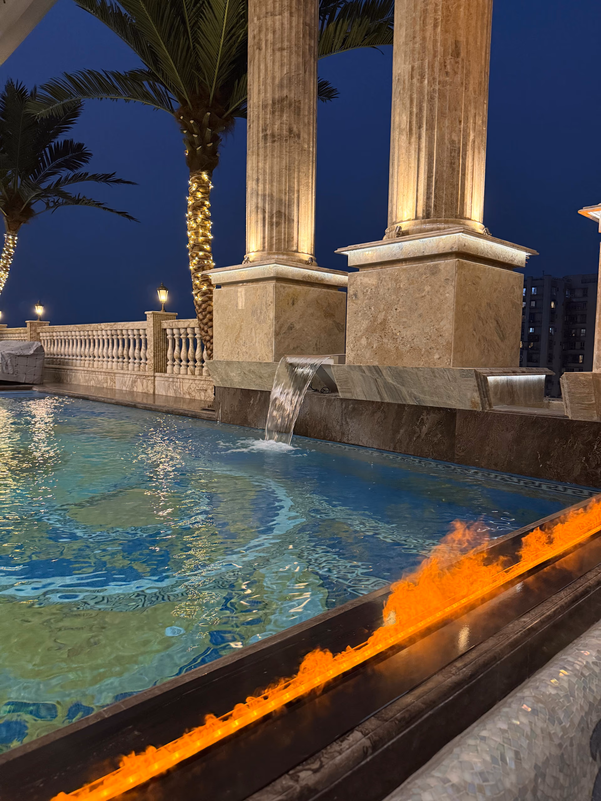Night view of a marble rooftop terrace with fluted stone columns, a linear fire feature, and an infinity-edge pool beside a balustrade.