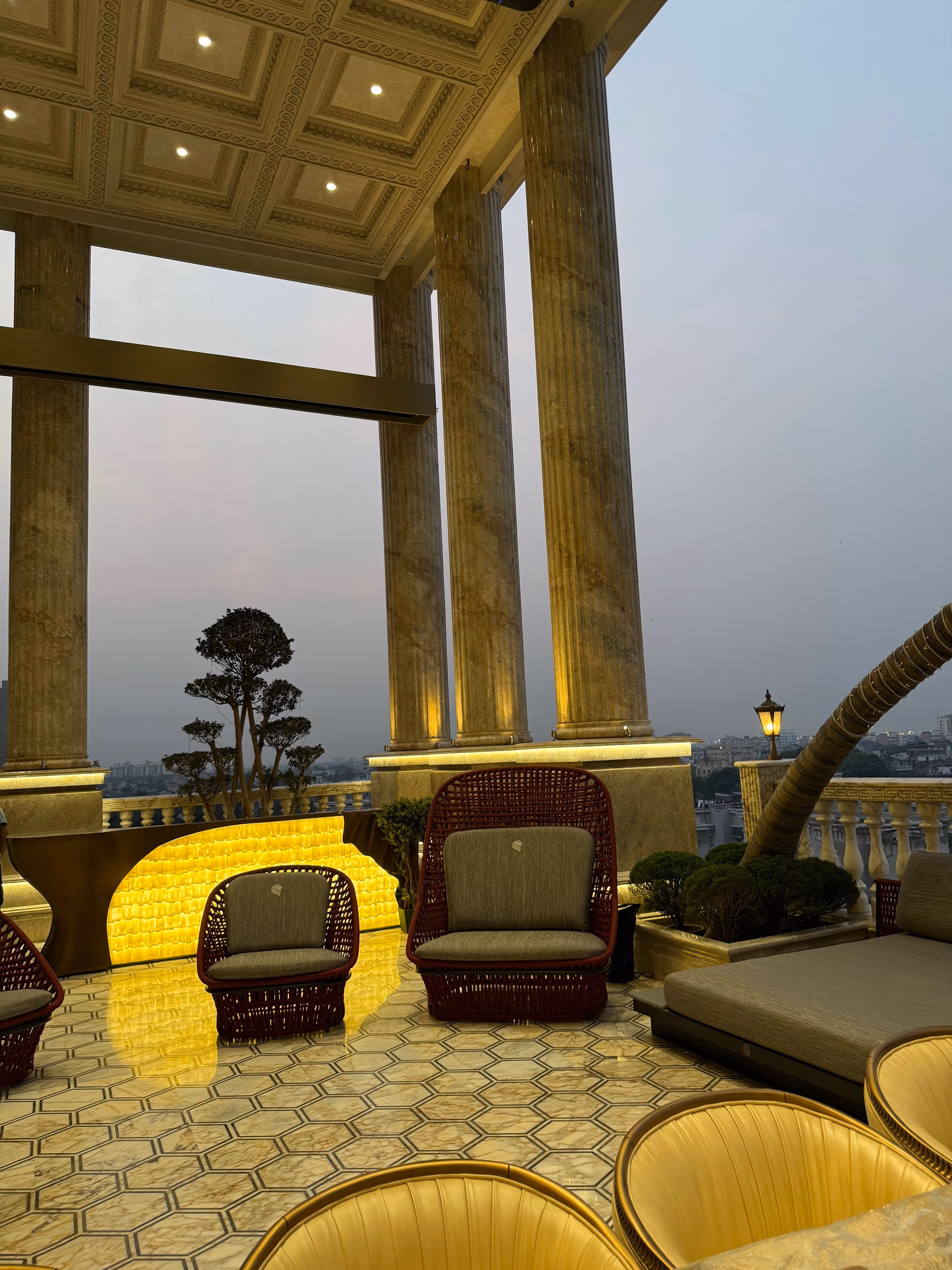 Terrace lounge with deep red woven barrel chairs, a daybed, and a hexagon marble floor under a coffered gold ceiling at dusk.