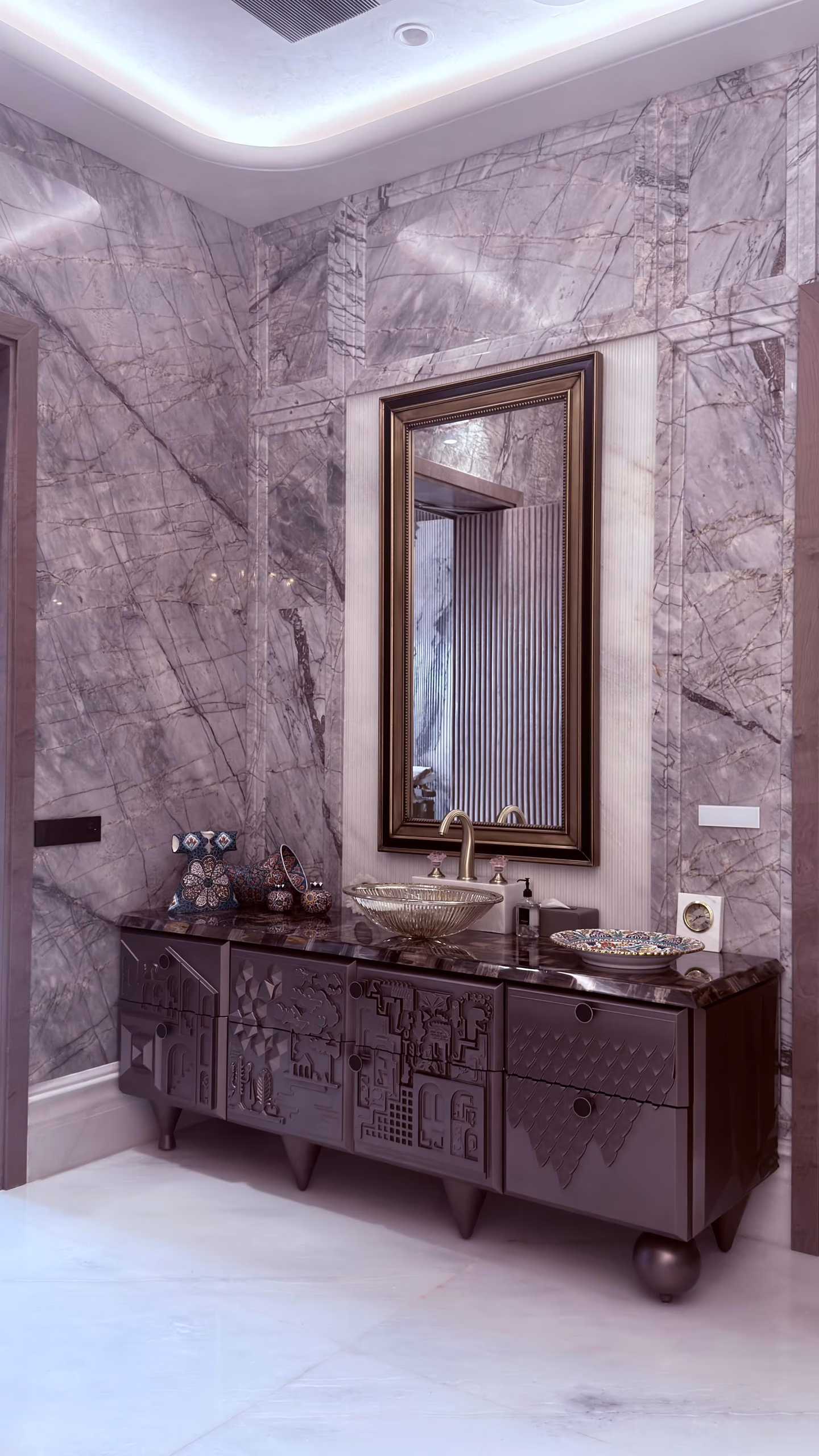 Mauve lacquer bathroom vanity with a sculpted cityscape relief front and stone top holding a glass vessel sink, framed mirror above on a grey marble wall.