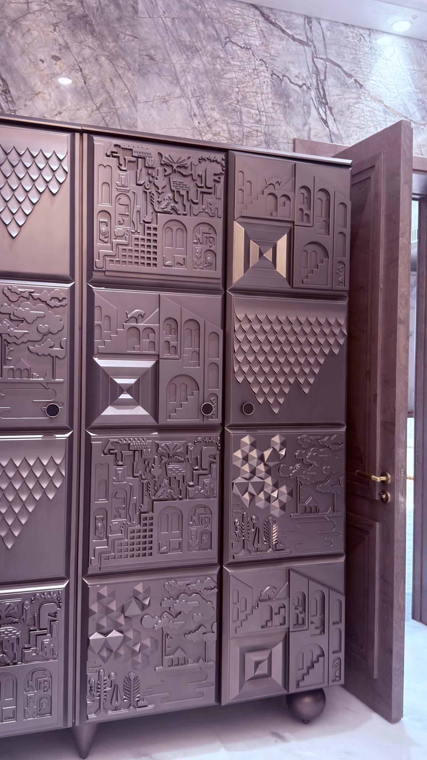 Close view of the vanity doors showing raised carved panels of houses, arches, trees and scale patterns in matte mauve, beside an open timber door.