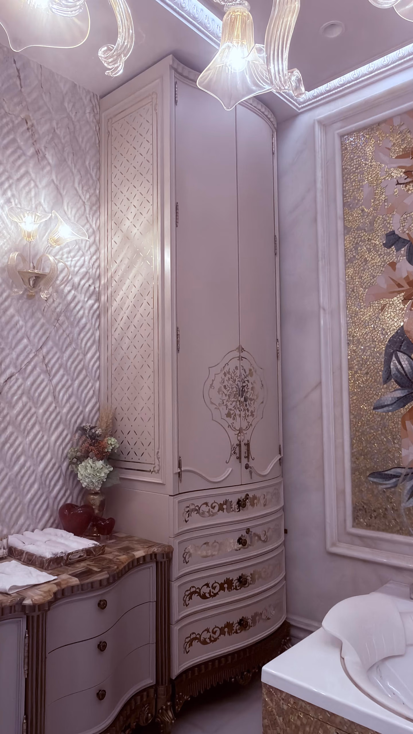 Tall white lacquer bathroom cabinet with gold foliate inlay and curved drawers, beside a marble-top vanity chest, under Murano glass lights and a mosaic panel.
