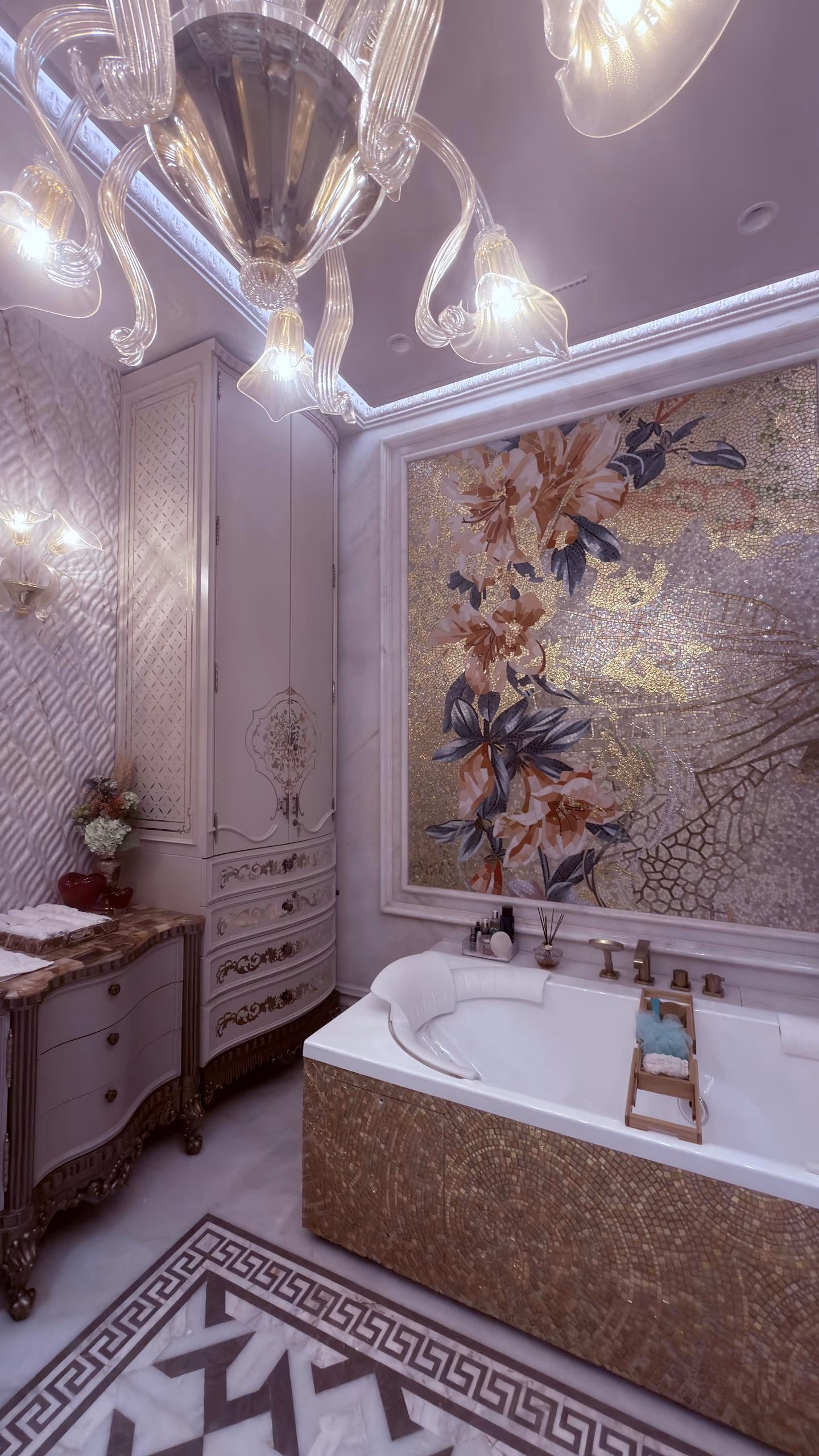 The white-and-gold cabinet and marble-top vanity chest in a full bathroom with a mosaic floral panel, gold-tiled tub surround and Murano glass chandelier.