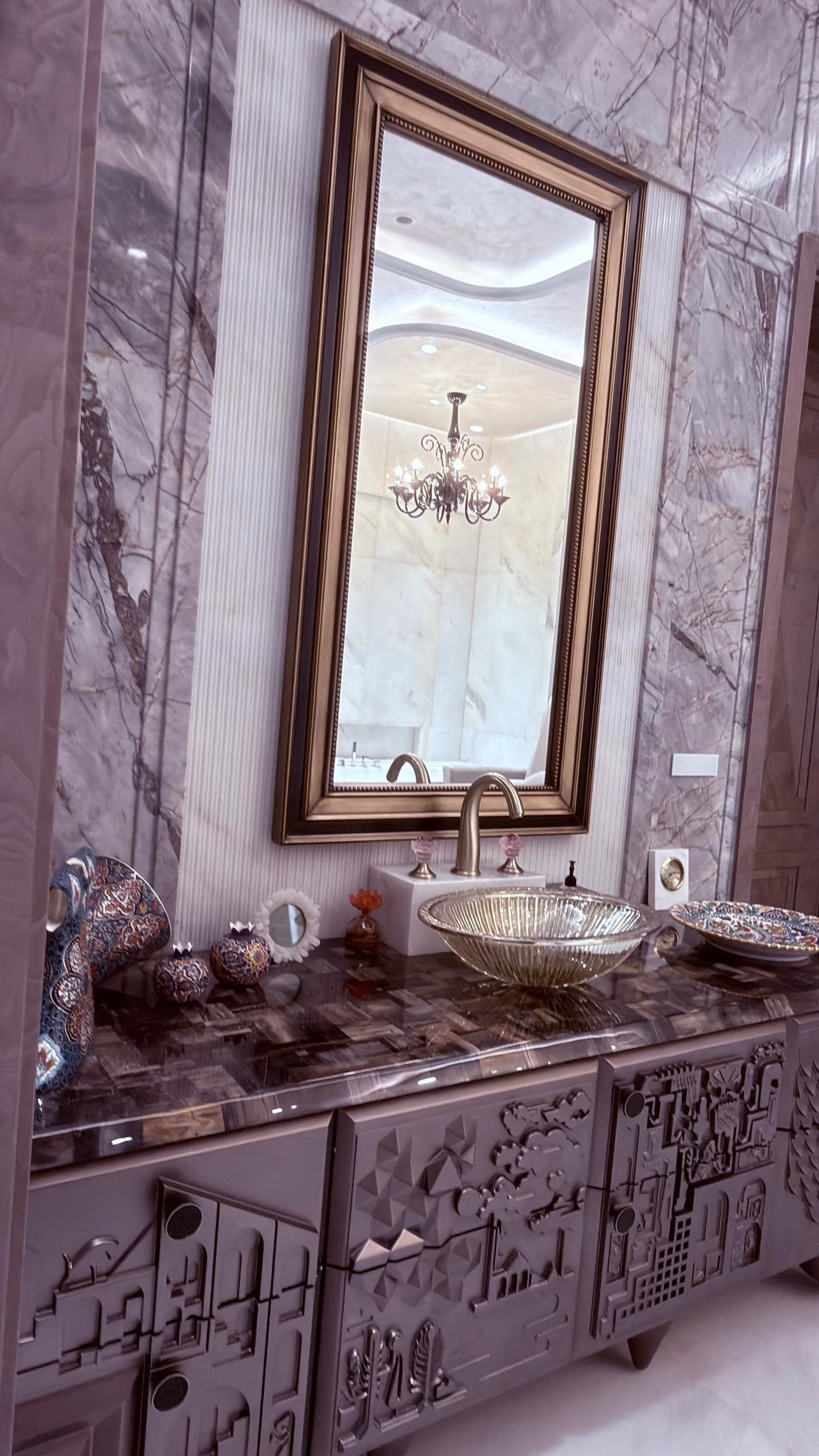 The pewter relief vanity seen from the side in a grey marble bathroom, its agate top and glass bowl below a tall gilt mirror reflecting a chandelier.