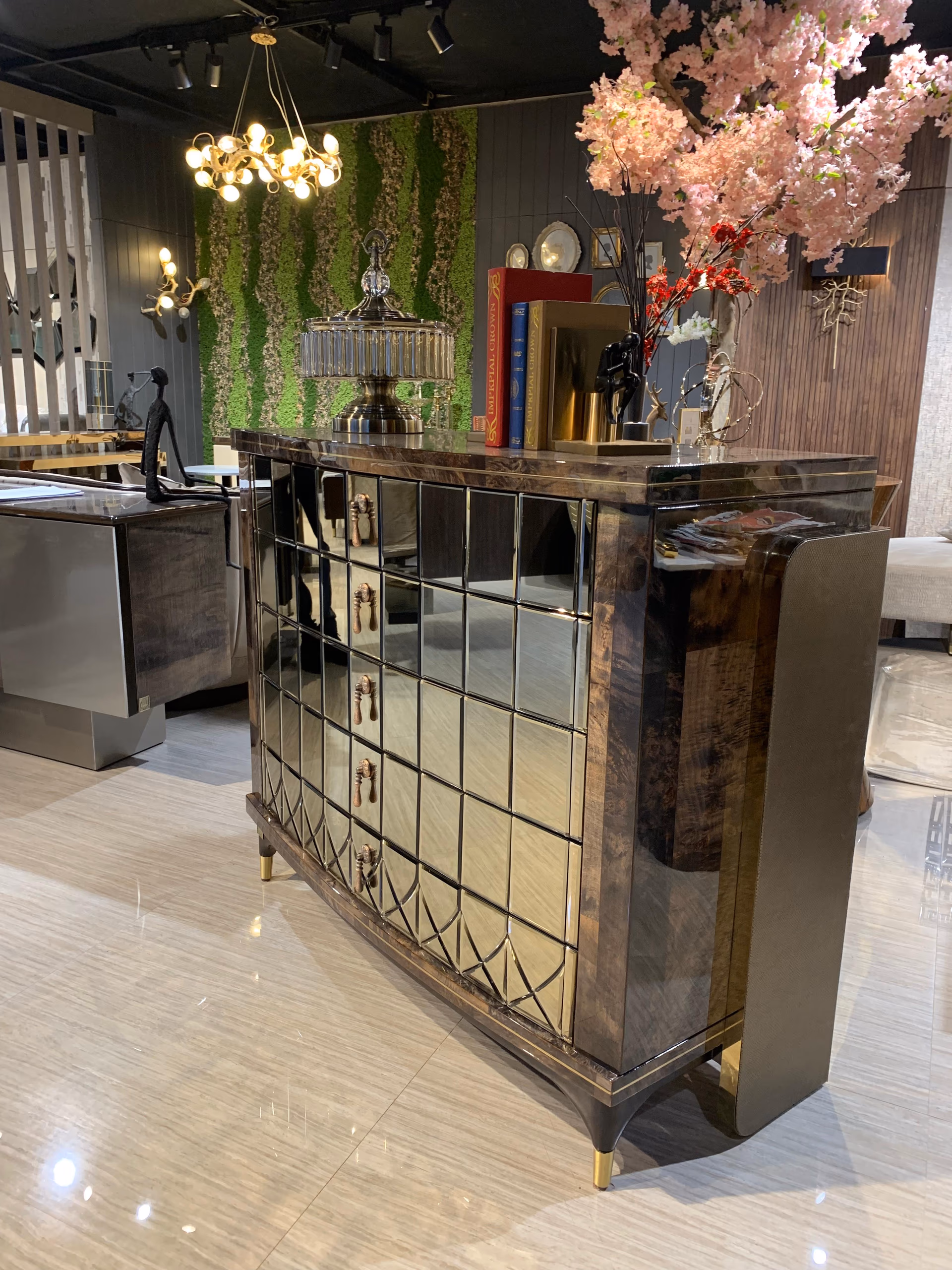 Angled view of the mirrored chest showing the burl-wood side, brass edge line and curved brass-tipped leg, styled with a glass urn and books on top.