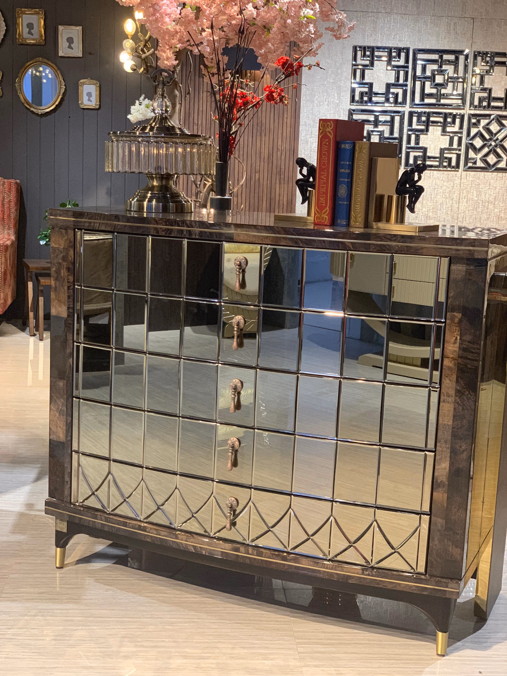 The mirrored chest of drawers styled with a lamp, books and blossoms, its bronze drop handles and diamond-cut lower drawer catching the light.