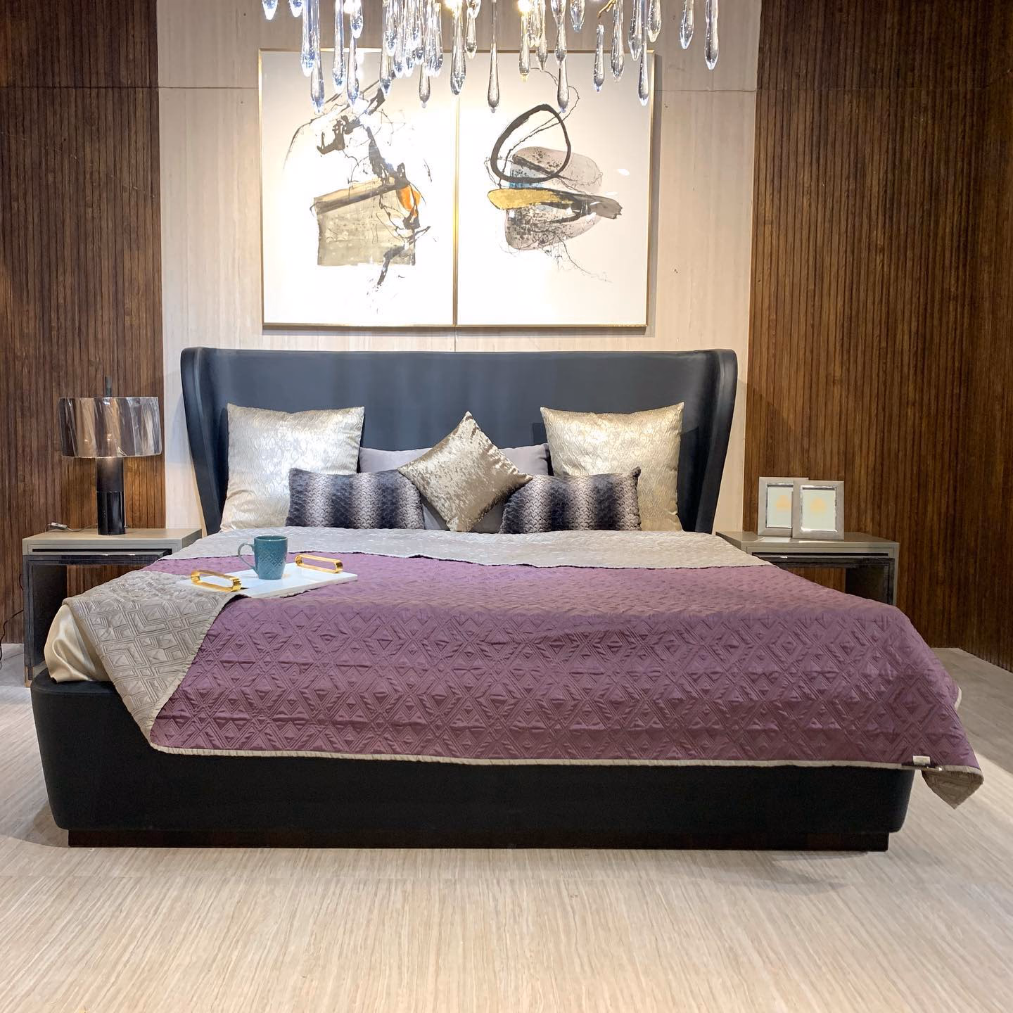 Black leather bed with a curved wingback headboard and a low platform base, styled under a crystal drop chandelier and a pair of abstract gold canvases in a fluted walnut bedroom.