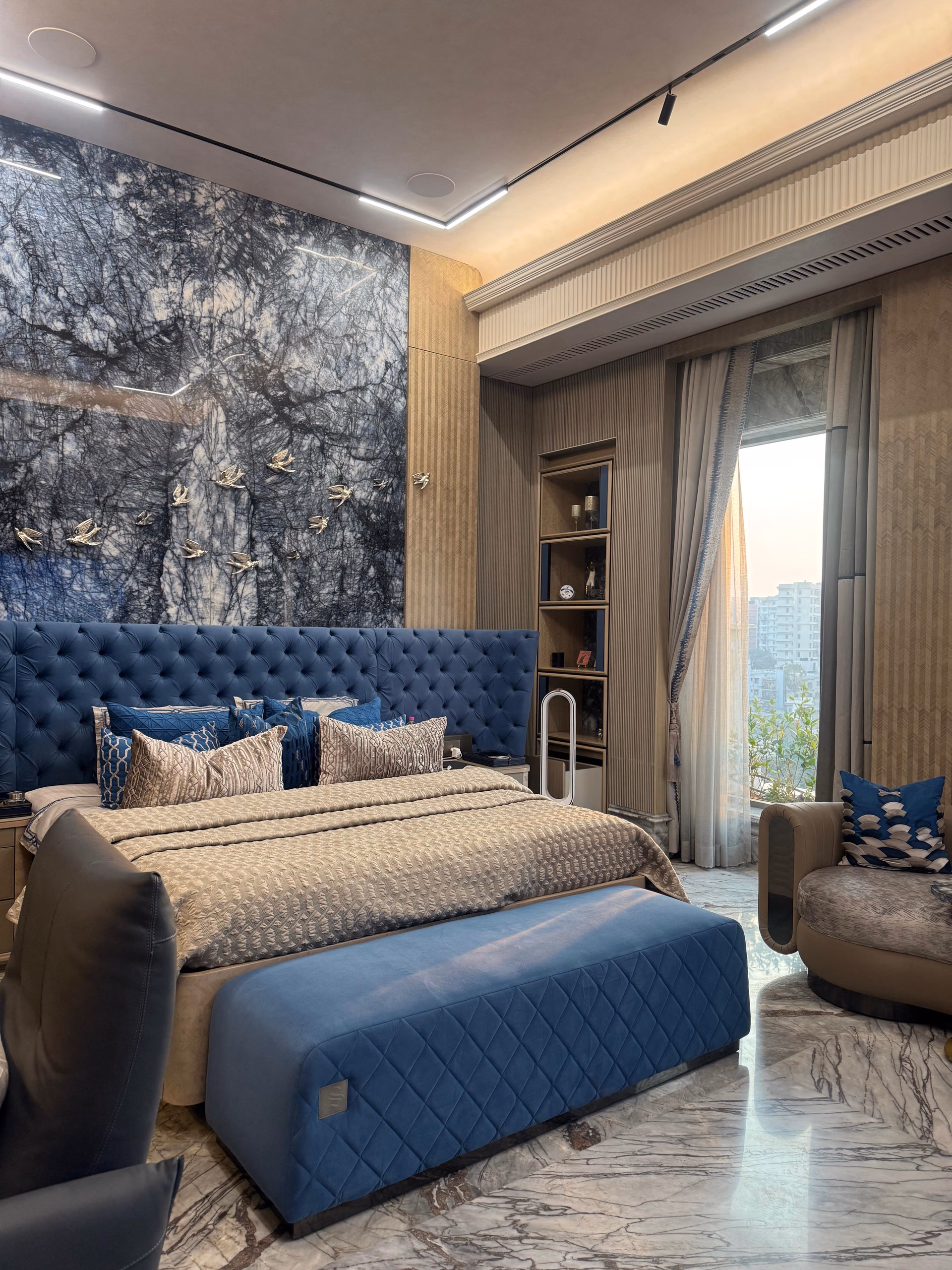 Full bedroom with a wide blue diamond-tufted headboard under a blue marble feature wall with brass bird inlays, a quilted blue bench at the foot, fluted wardrobes and a curved lounge chair by the window.