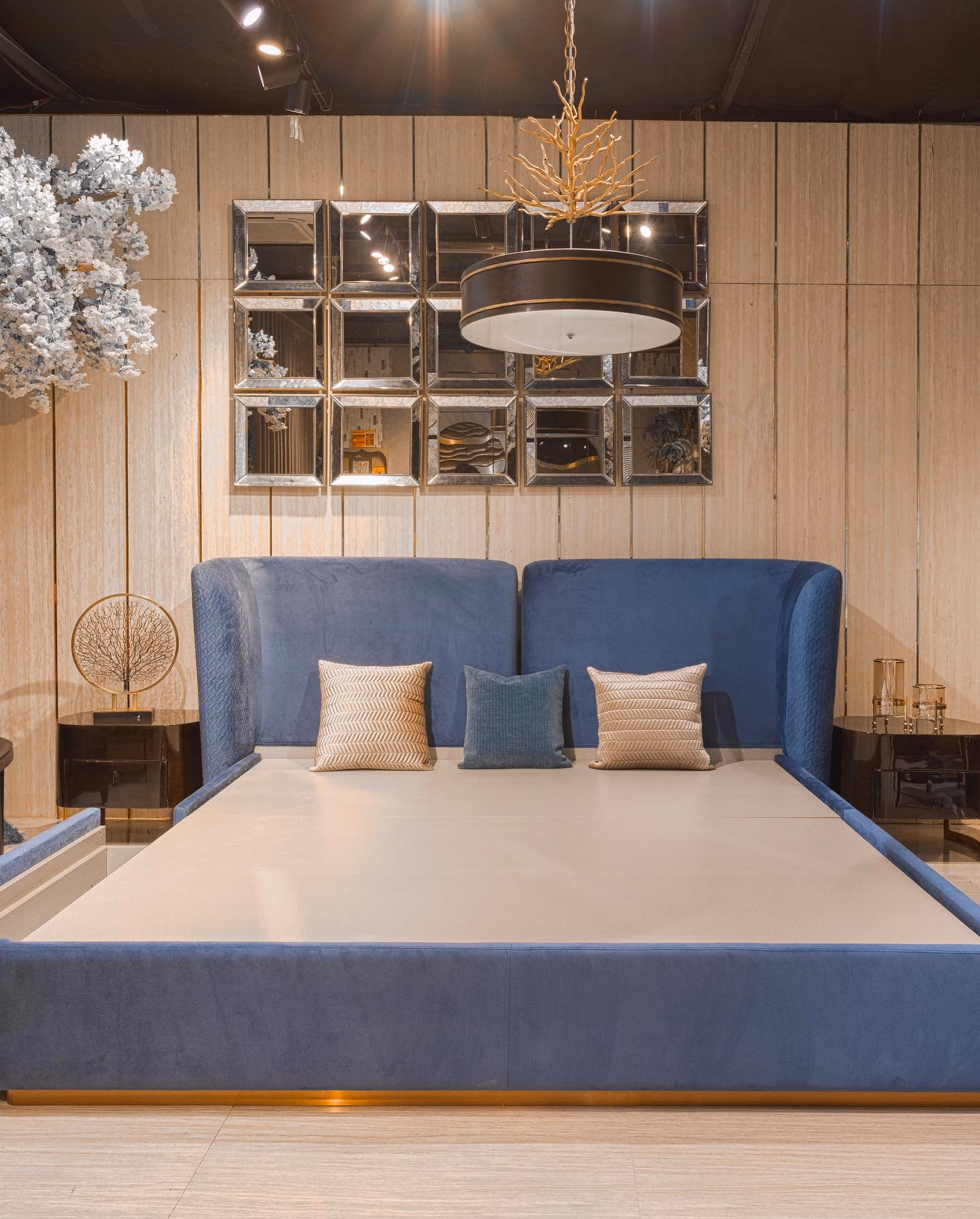Blue velvet platform bed with a two-part winged headboard and a brass-lit plinth base, shown under a bevelled square mirror panel set and a black drum pendant light.