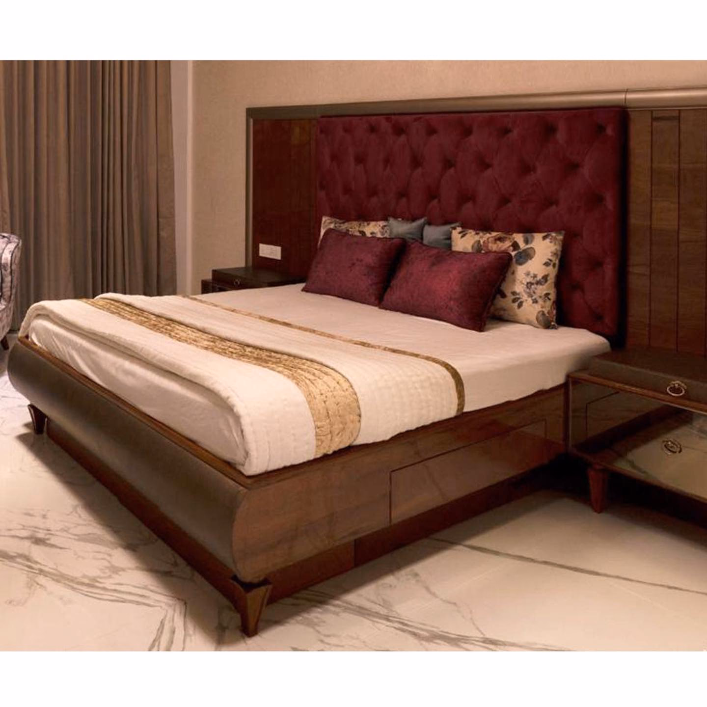 King bed with a tall burgundy diamond-tufted headboard set into a walnut panel wall, curved walnut storage base with drawers and a matching bedside table on a marble floor.