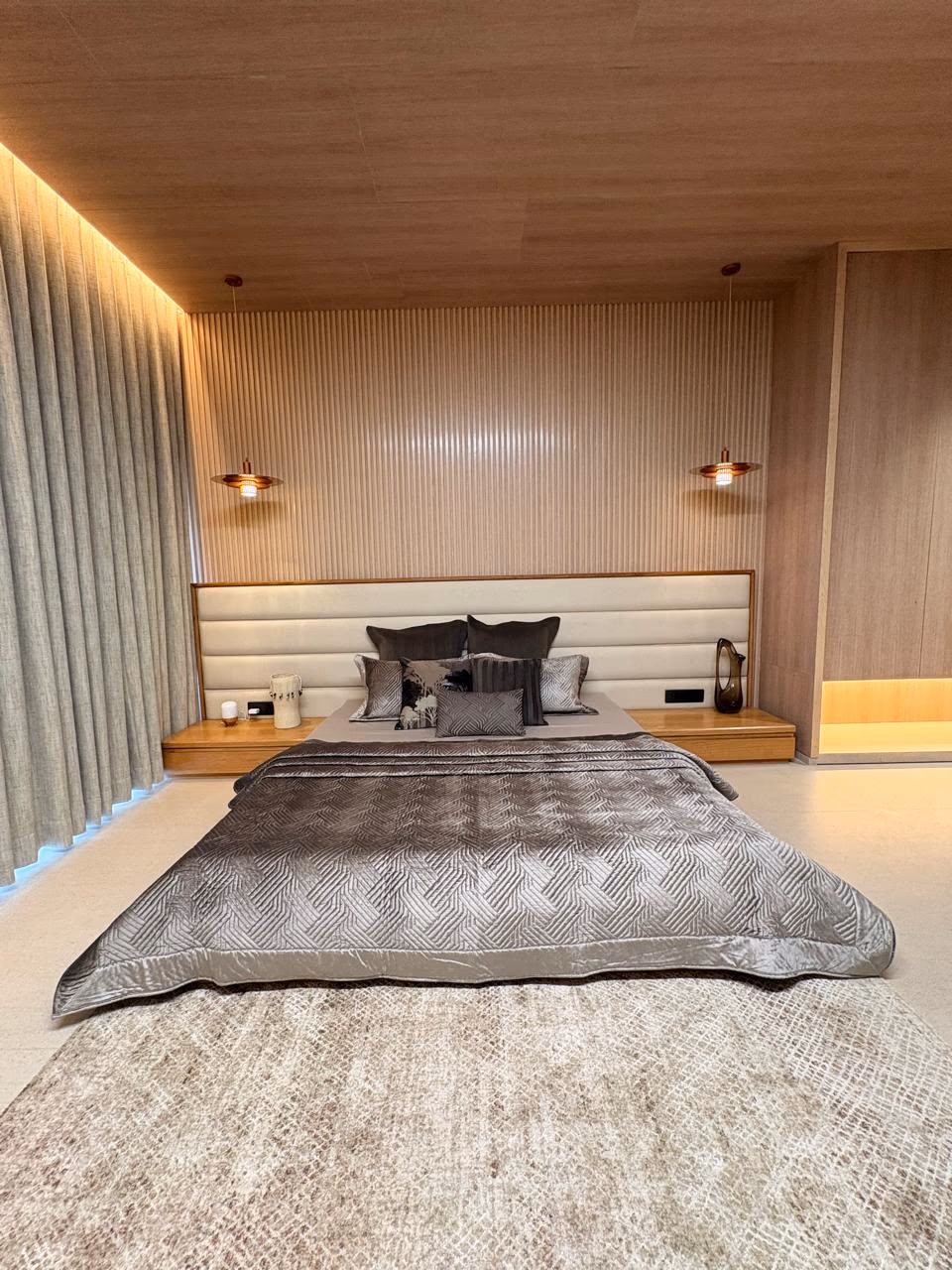 Wide ivory channel-tufted headboard against a ribbed wood-paneled wall, floating nightstands, and a silver quilted bedcover, flanked by drop pendant lights.