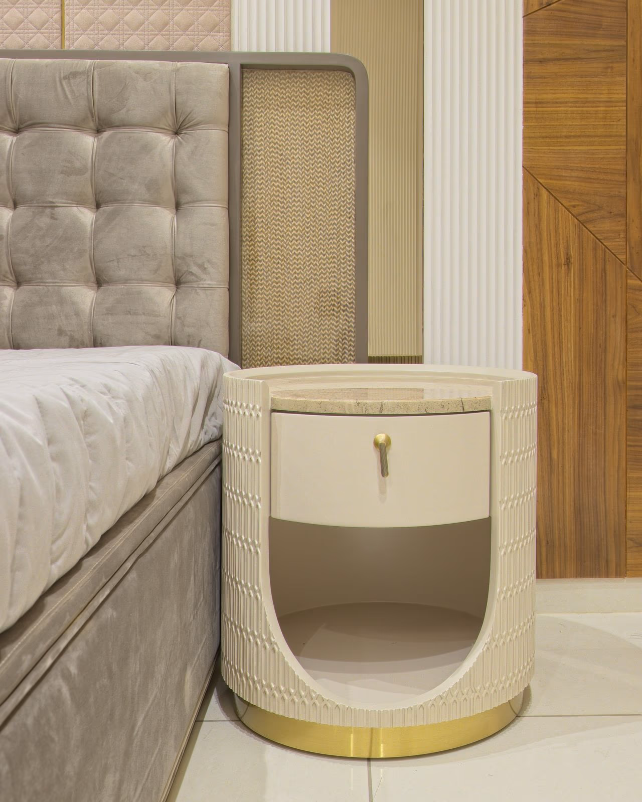 Round cream bedside table with diamond-carved sides and a brass ring base beside a grey tufted velvet bed.