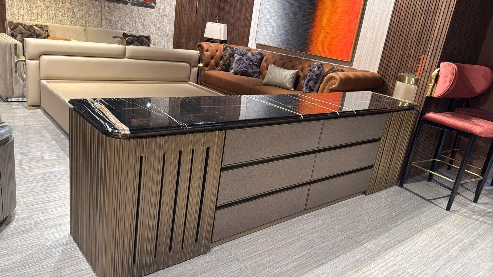 Low six-drawer dresser with a black marble top, fabric drawer fronts and a fluted champagne-gold metal base, shown in a showroom beside a tan leather sofa.