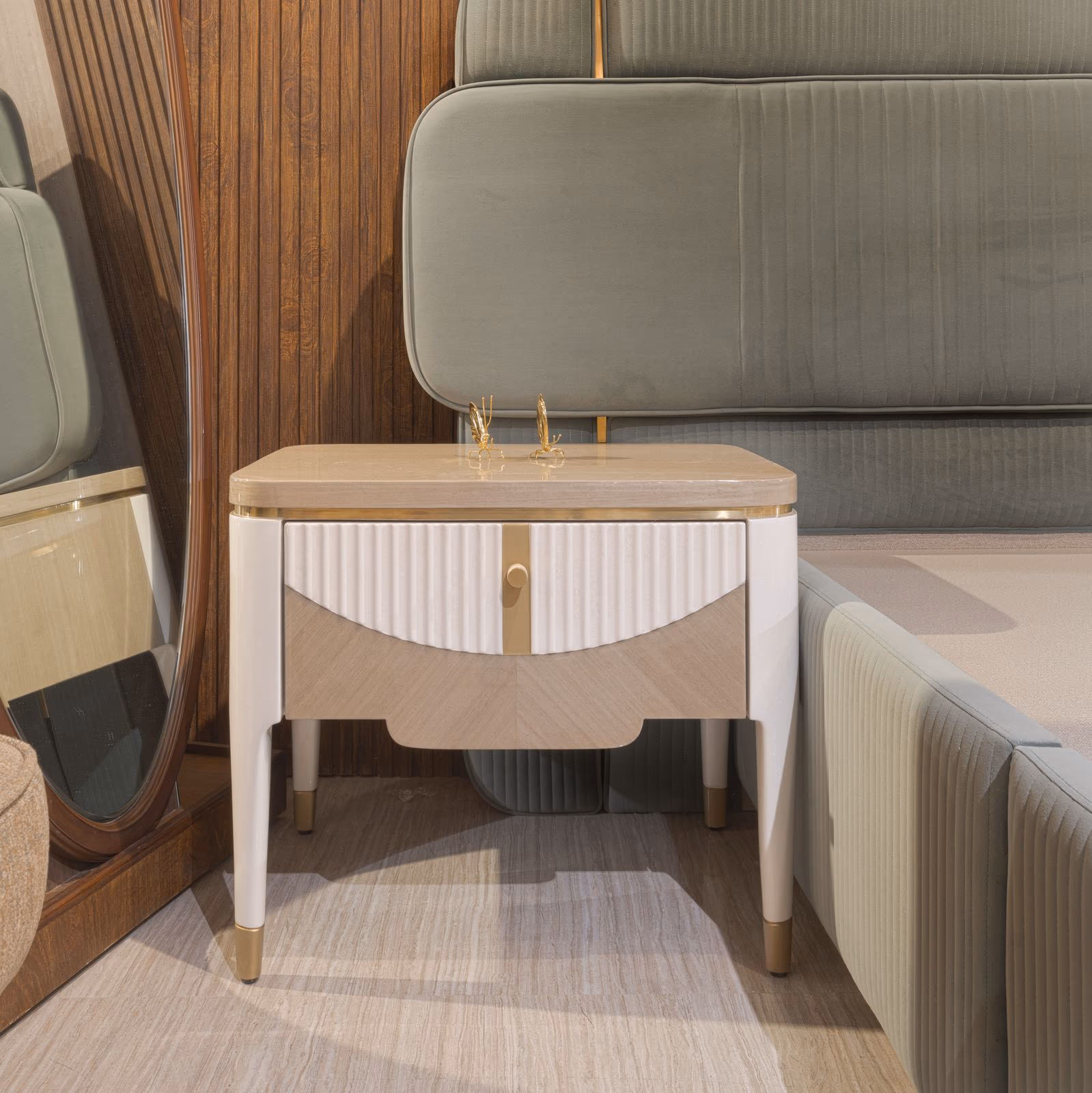 Ivory lacquer bedside table with a fluted cabinet door, brass edging, tapered legs, and a stone-look top, staged beside a green velvet headboard.