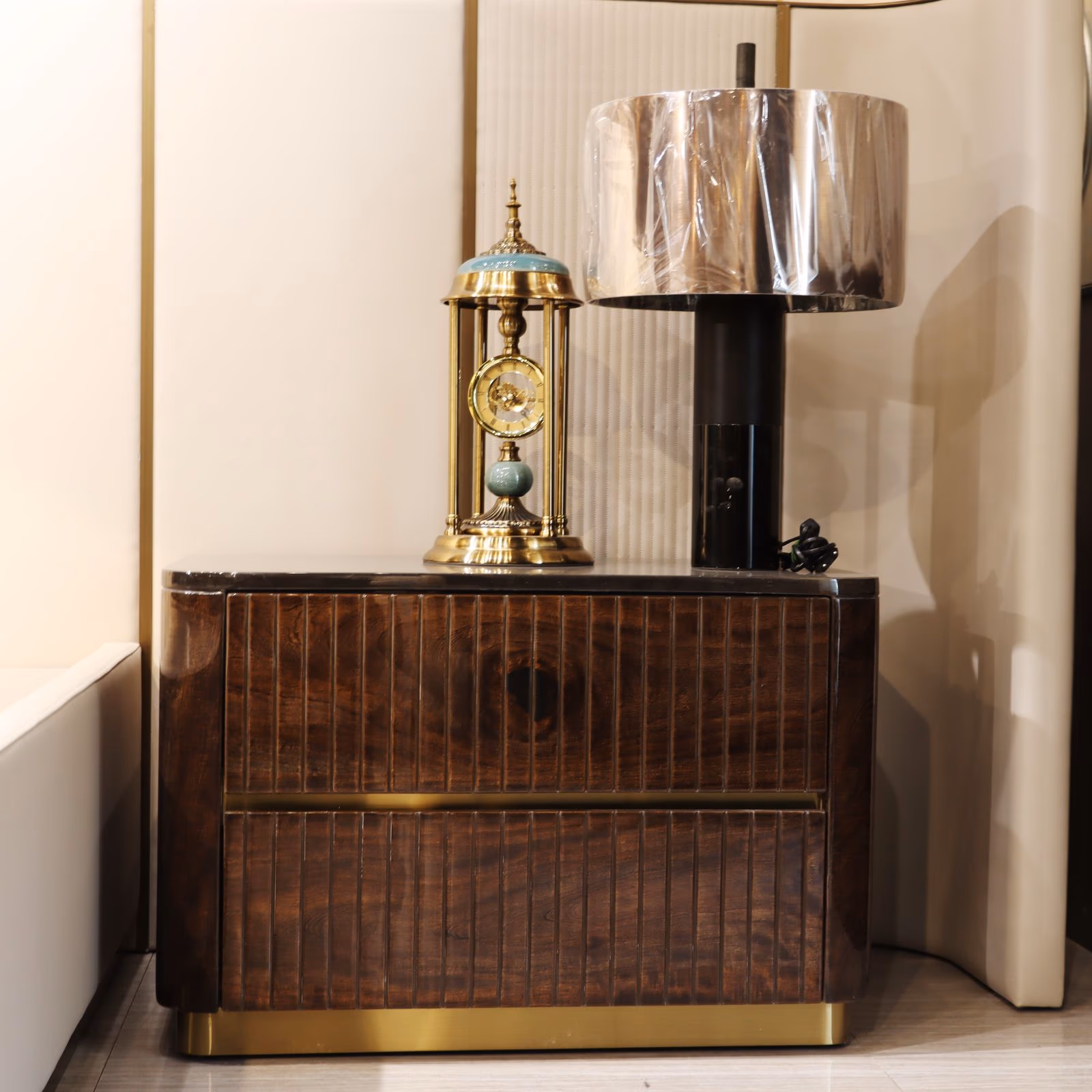 Dark walnut two-drawer nightstand with vertical fluted panels and a brass base, topped with a gold clock and lamp.