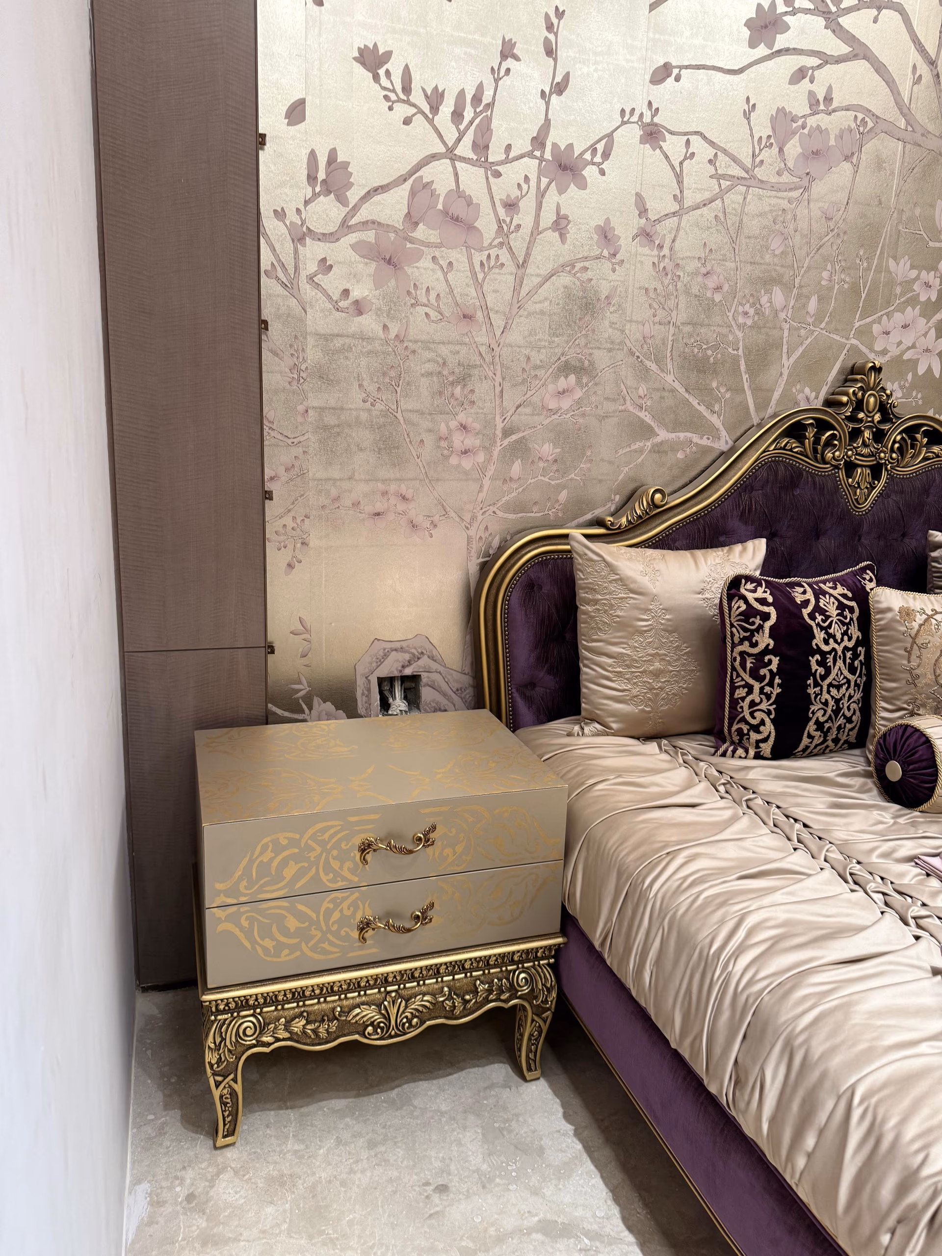 Gold gilt stencilled bedside table with carved legs beside a purple upholstered bed and chinoiserie wall
