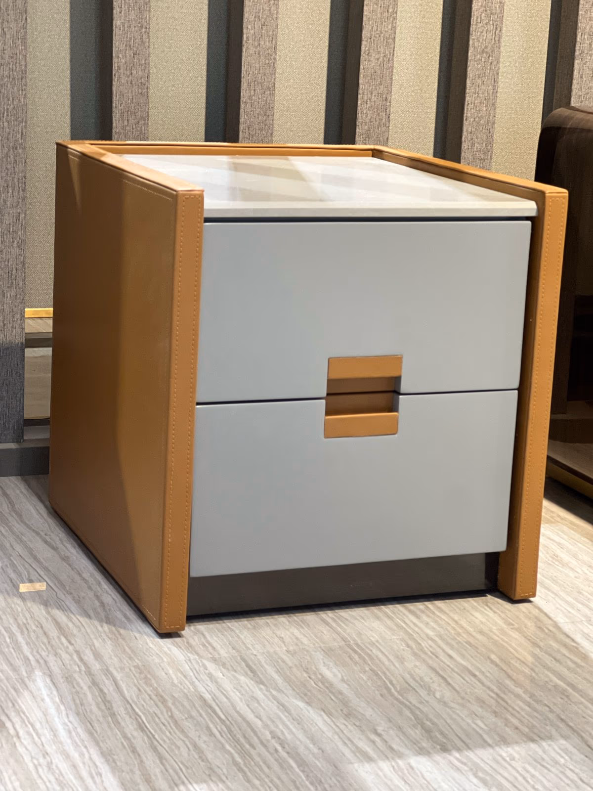 Grey lacquered two-drawer nightstand framed in tan leather with a square brass handle and white marble top.