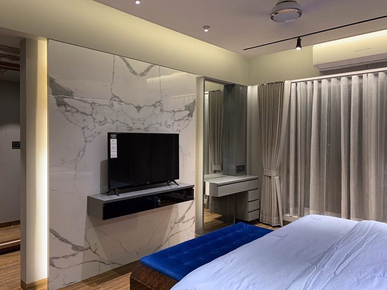 Bedroom television wall clad in book-matched white marble with a slim floating grey console below the screen, a mirrored dressing niche alongside and a blue velvet bench at the foot of the bed.