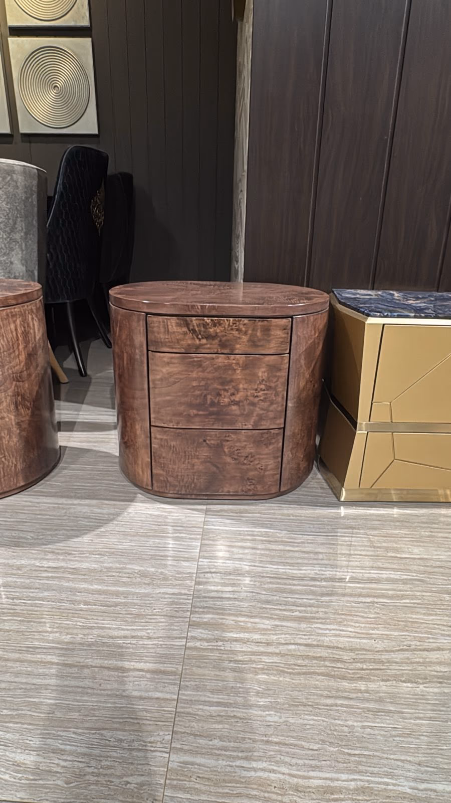 Cylindrical bedside table in rich brown burl wood veneer with three stacked drawers, shown in a furniture showroom.