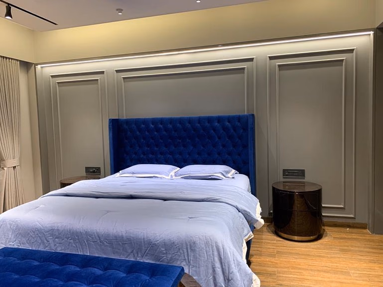 The royal blue tufted wingback bed dressed in pale blue linen in a grey moulded-panel bedroom, with a blue velvet bench at the foot and a dark drum bedside table.