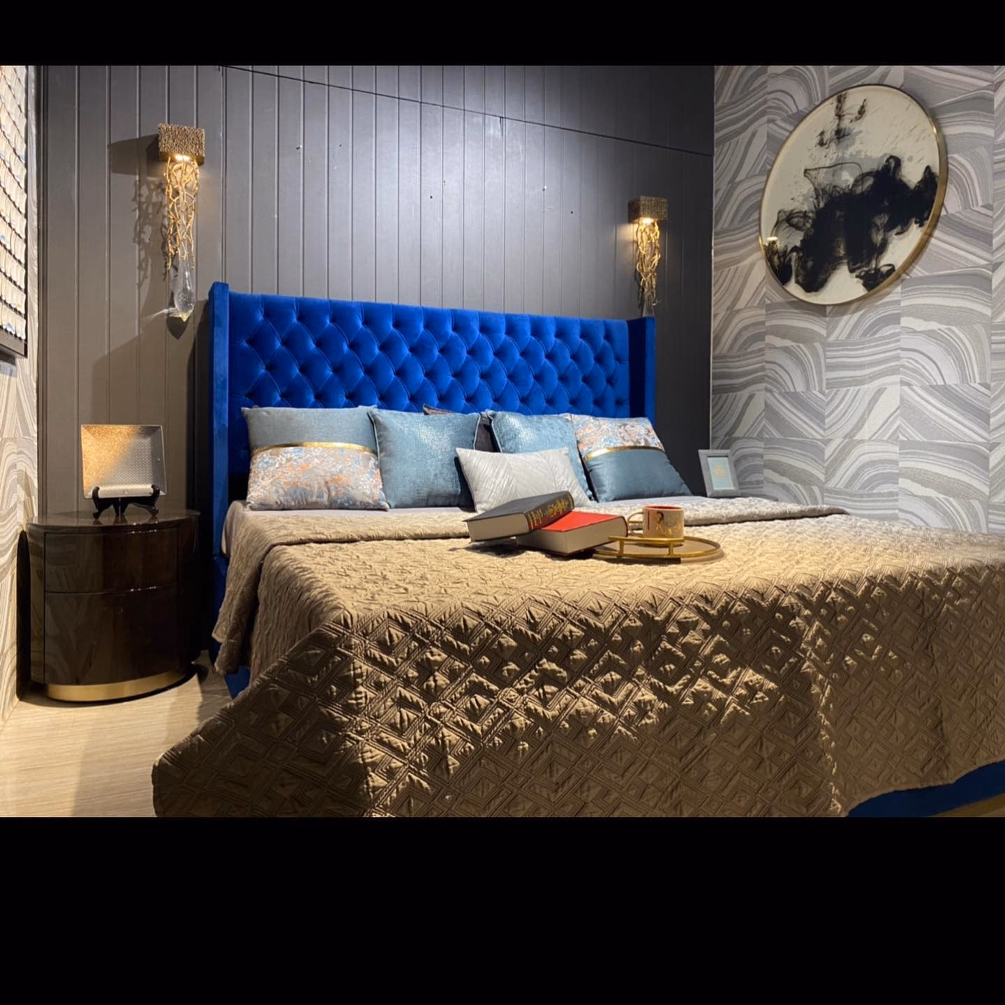 Royal blue velvet bed with a wide diamond-tufted wingback headboard against a grey slat wall, styled with a burl-wood round nightstand and gold crystal wall lights.