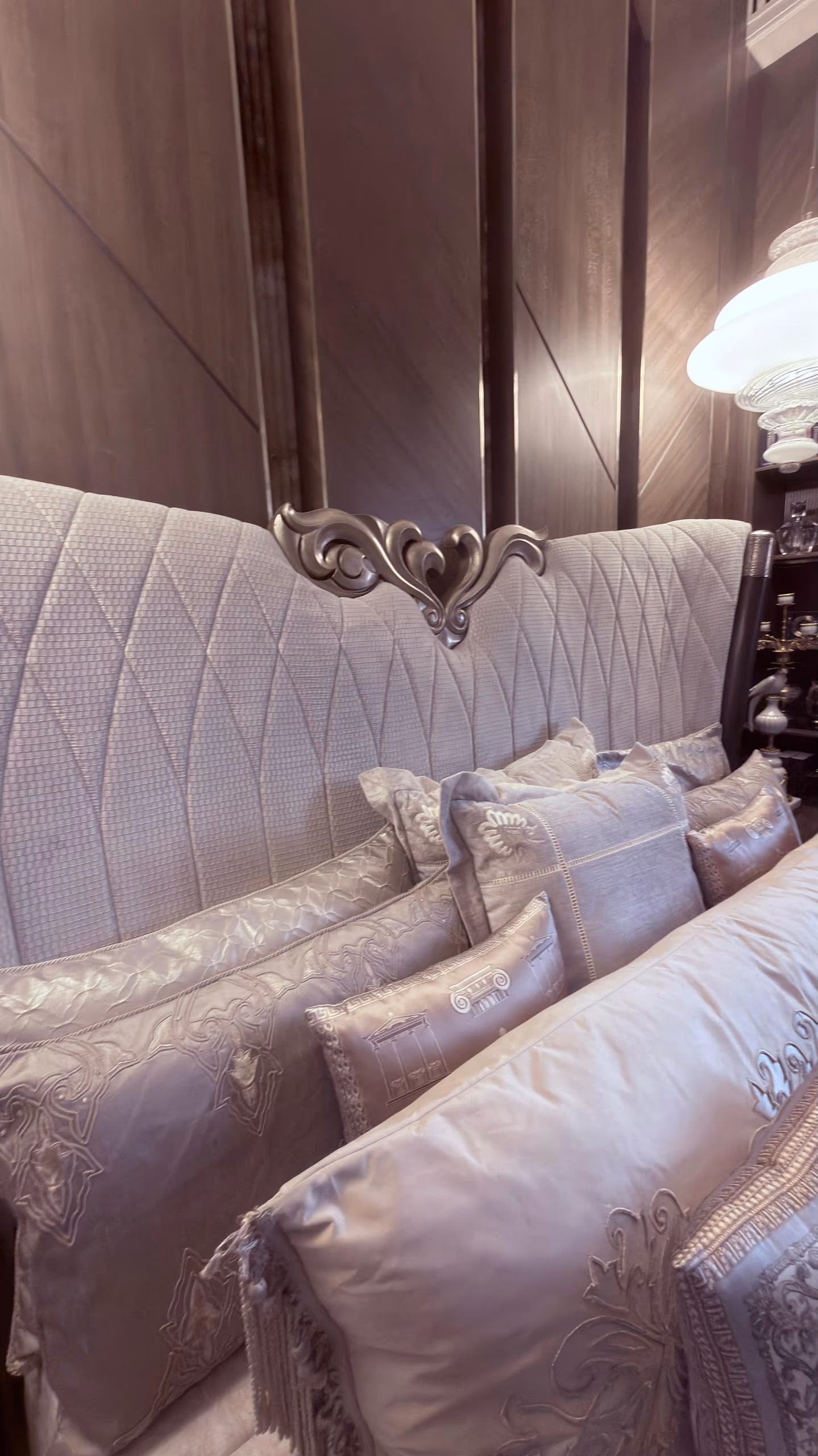 Tall channel-quilted grey satin bed headboard topped with a carved silver foliate crest, layered with silk pillows and an embroidered bolster in a panelled room.