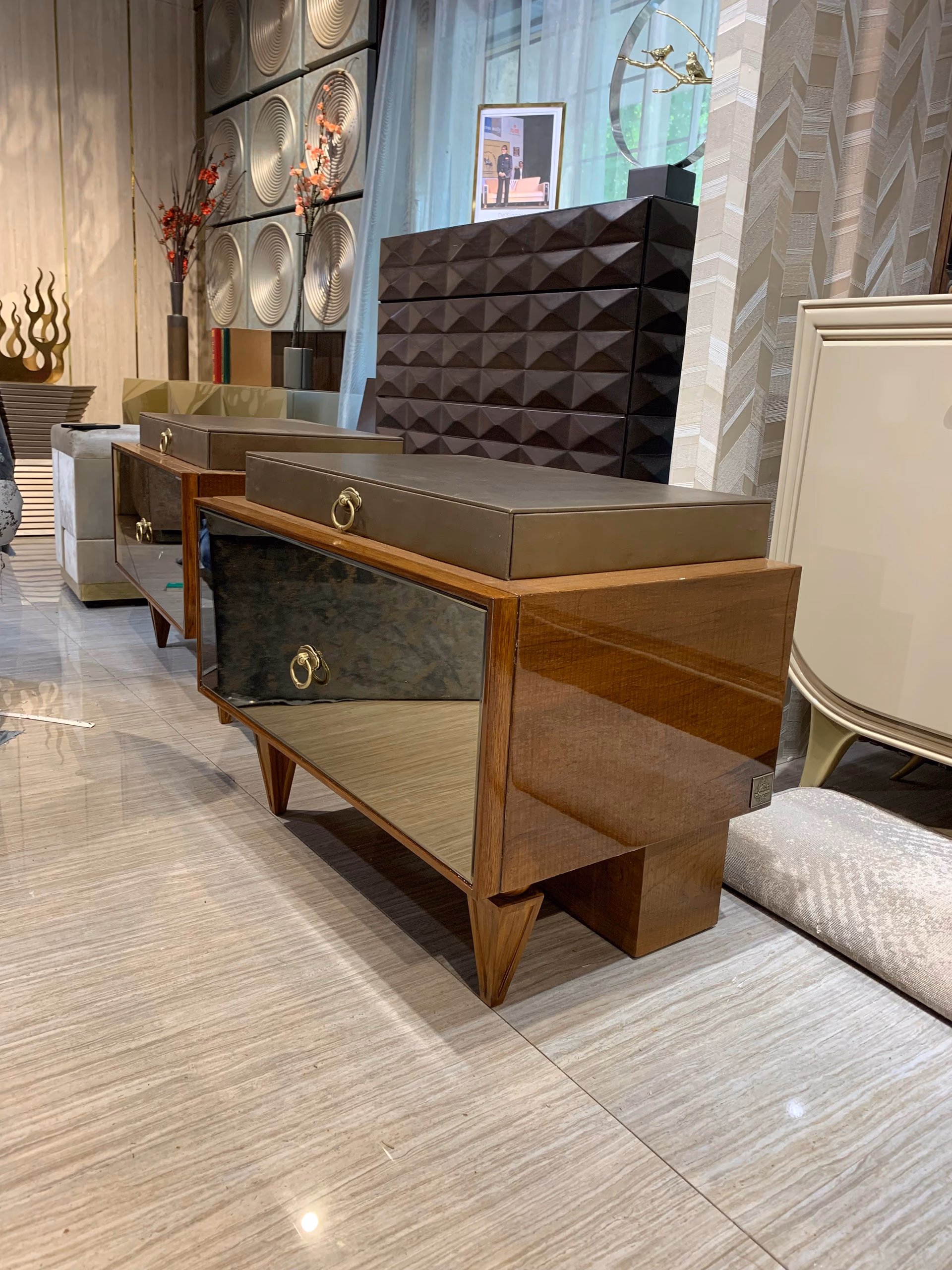 Bedside table in glossy walnut with a bronze-mirror drawer front, a brass ring pull and sharp tapered legs, part of a row of nightstands in a showroom.