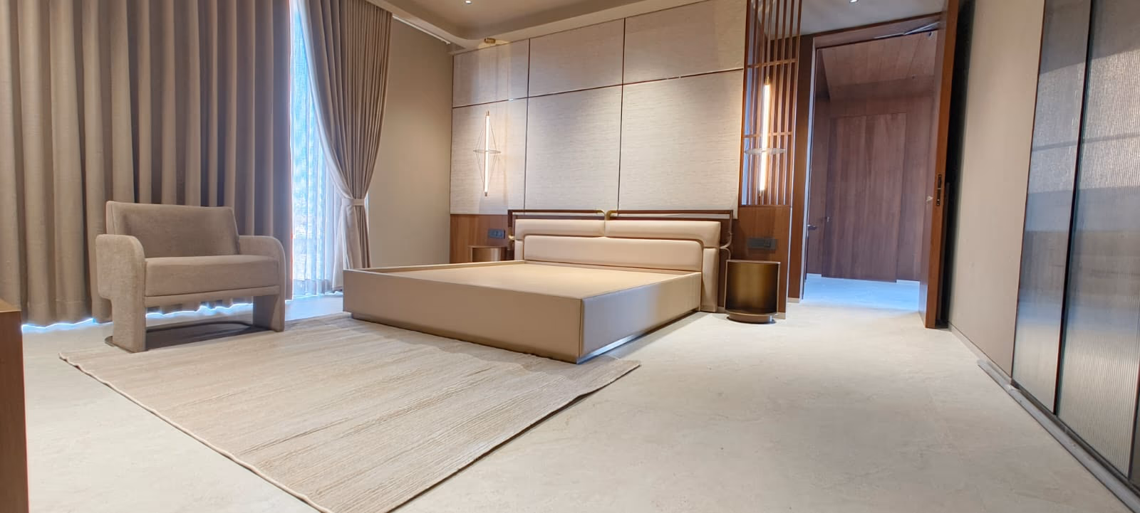 Low platform bed with a padded ivory headboard and waterfall base, shown in a full room with an armchair, rug, and open wardrobe.