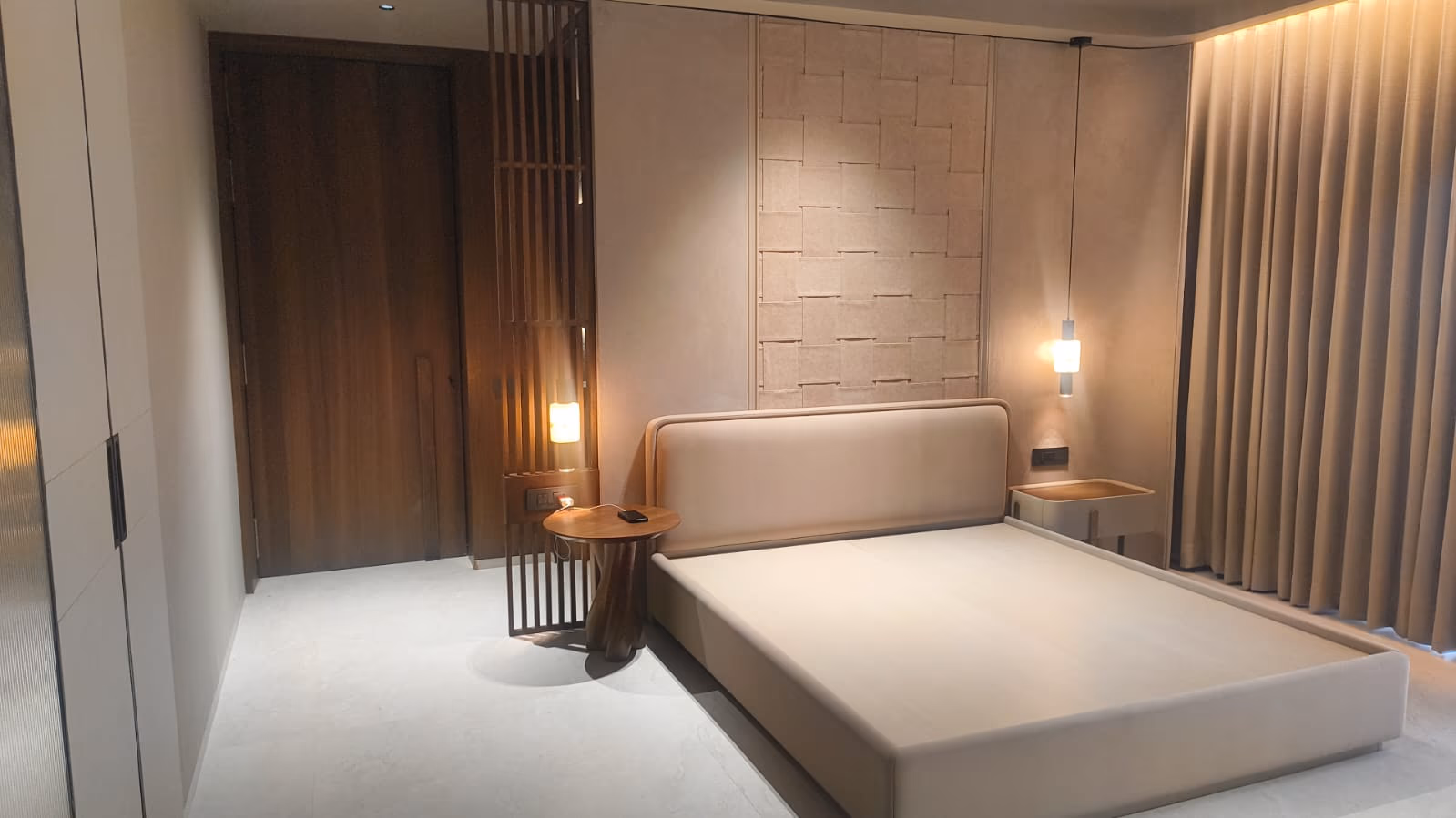 Wide view of a beige bedroom showing the woven leather headboard wall, a round wood side table, wardrobe, and wood entry door.