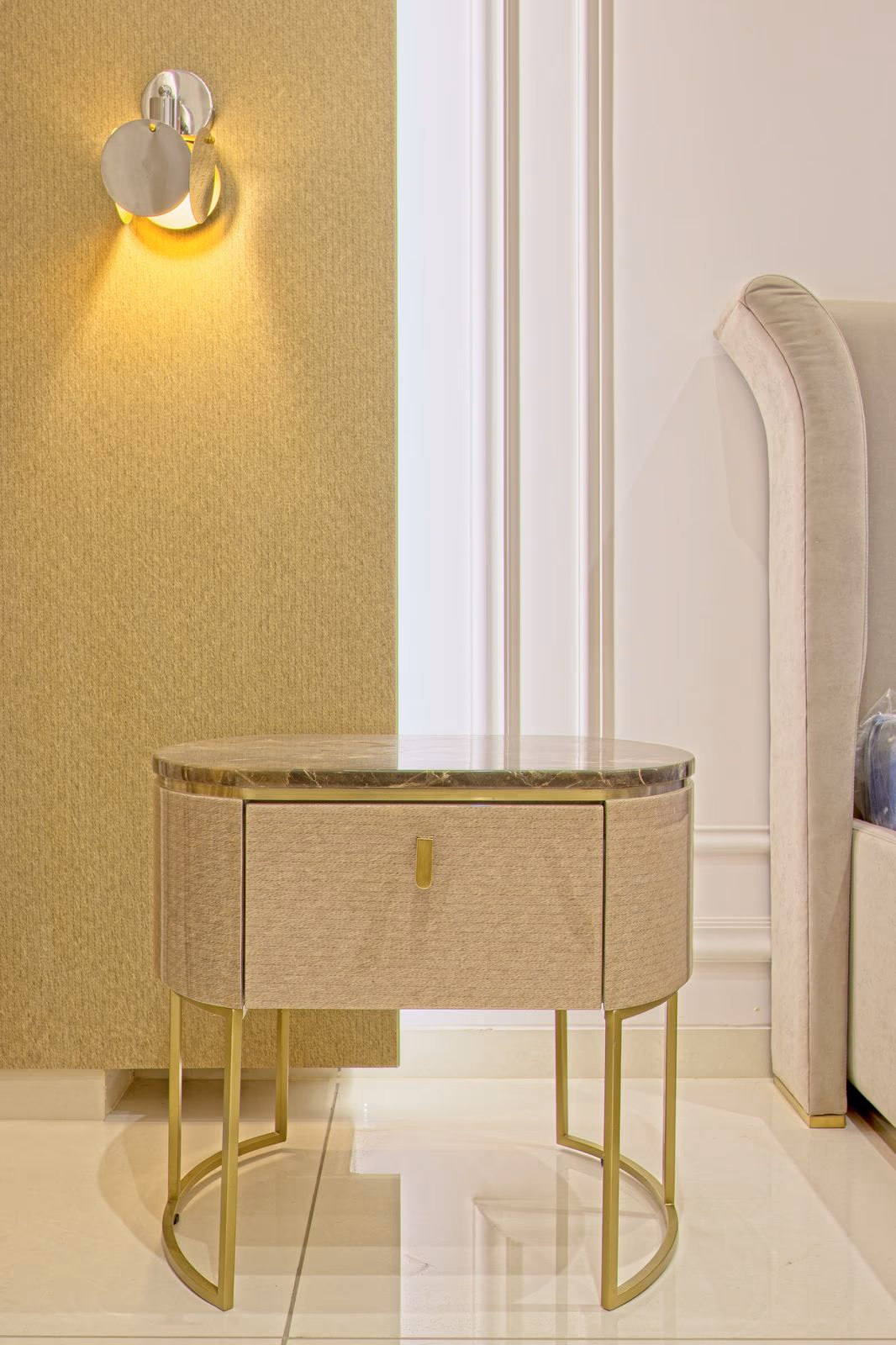 Oval bedside table with woven fabric front, a stone top, and slim brass tube legs beside a beige upholstered bed.