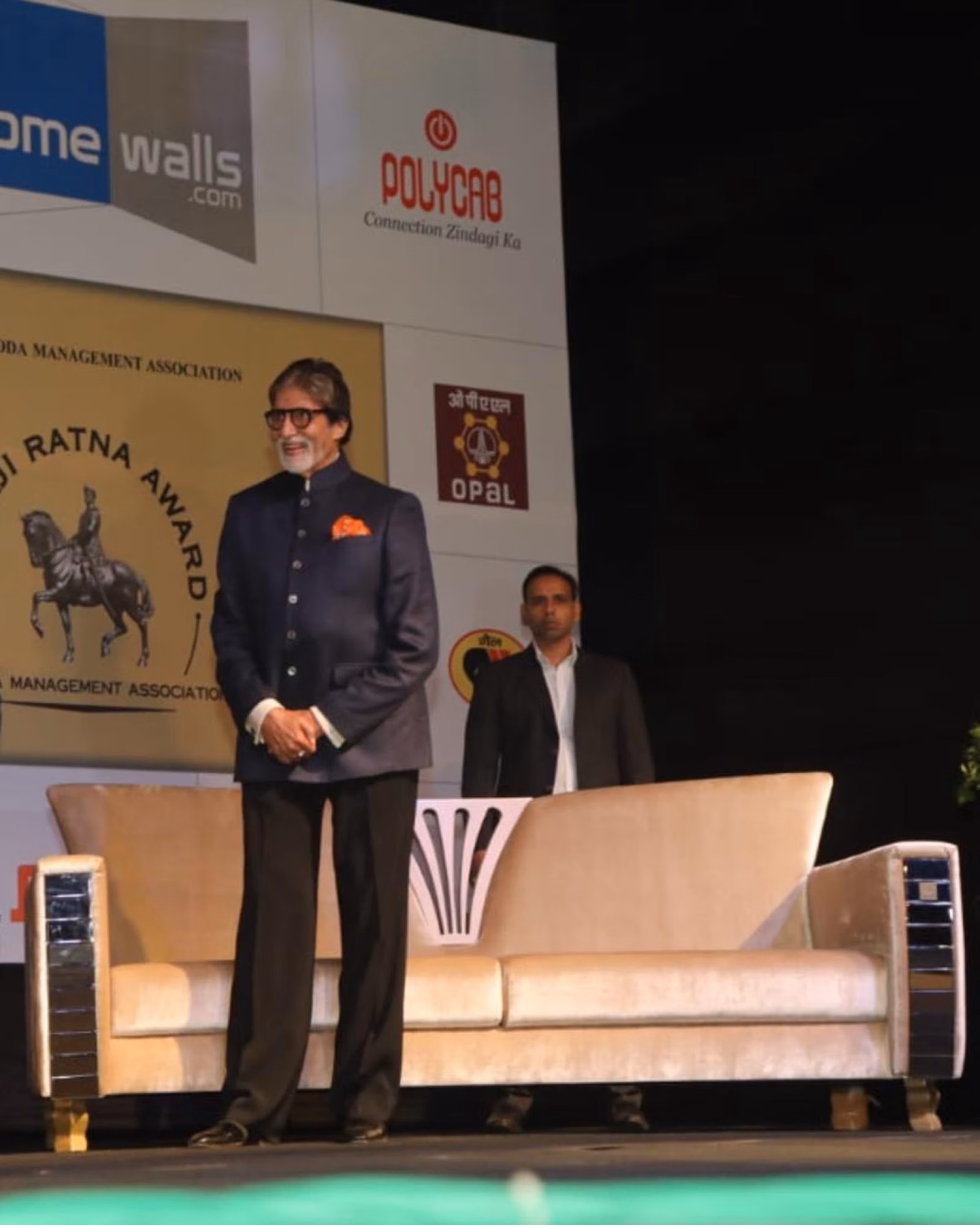 Amitabh Bachchan on stage beside a cream Designatore sofa with mirrored-and-brass sides.