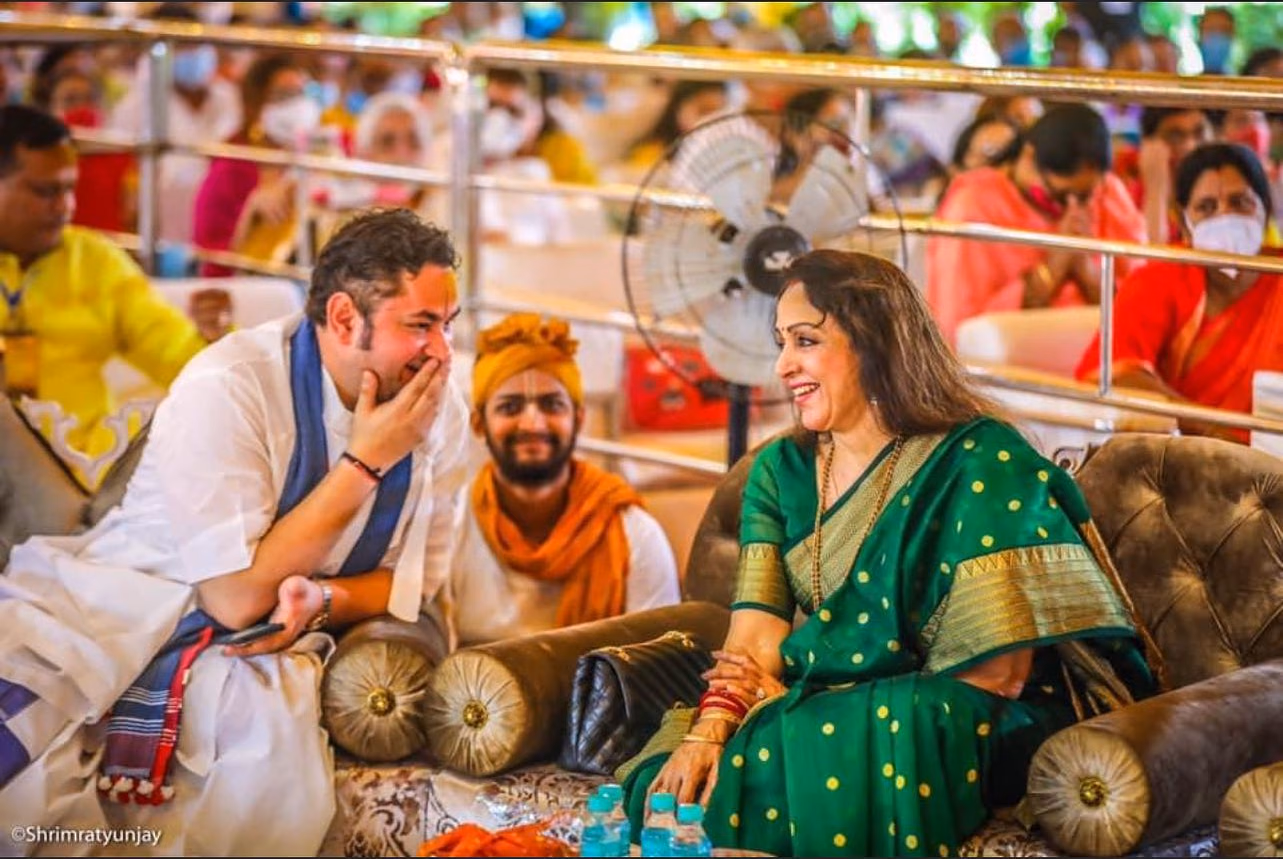 Hema Malini in a green silk saree laughing with Shri Pundrik Ji Goswami on a deep-buttoned Designatore sofa.