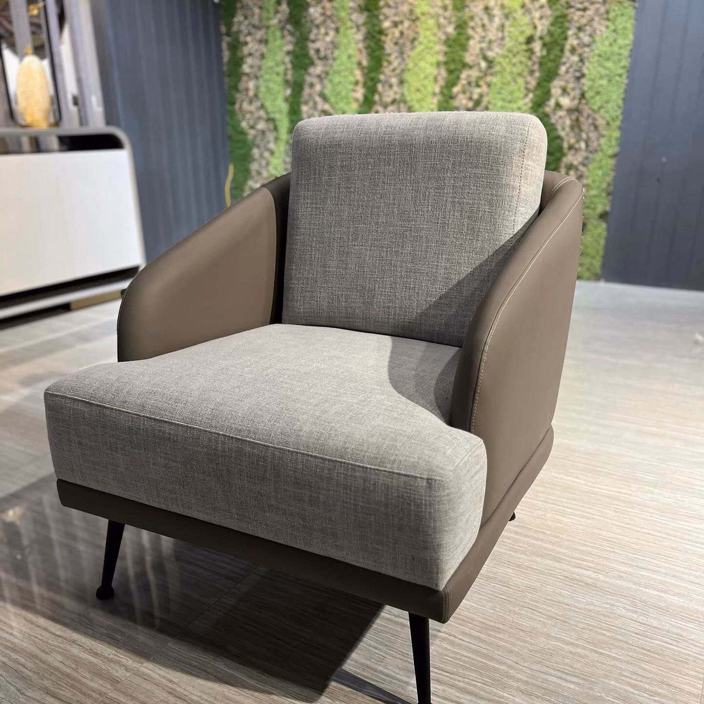 Curved-arm accent chair in grey fabric and taupe leather on slim black metal legs against a green living wall.