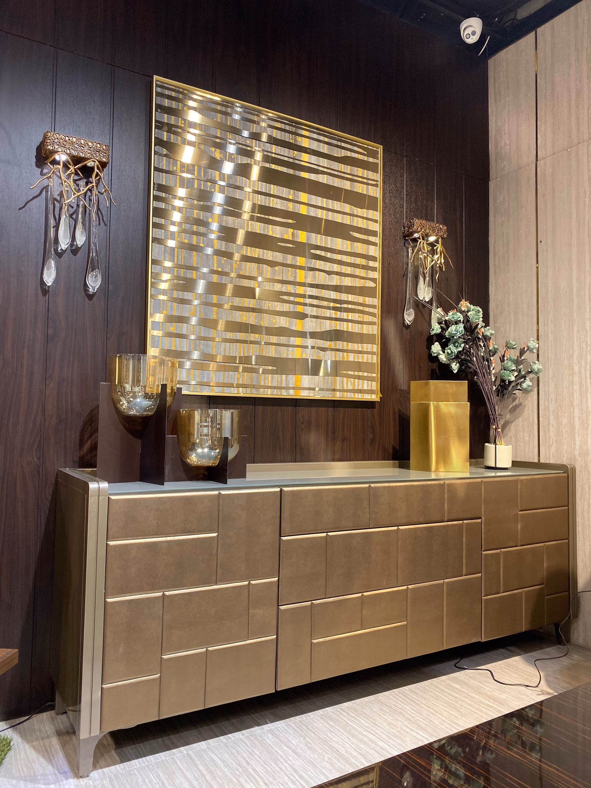 Long champagne sideboard with a three-dimensional block-textured leather front, silver metal trim and a glass top, set below framed gold art on a dark wall.
