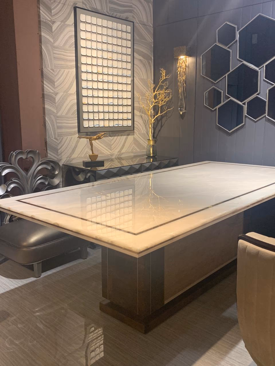 Rectangular dining table with a cream marble top bordered by a thin brass inlay line, carried on a wide two-tone wood pedestal base on a grey floor.