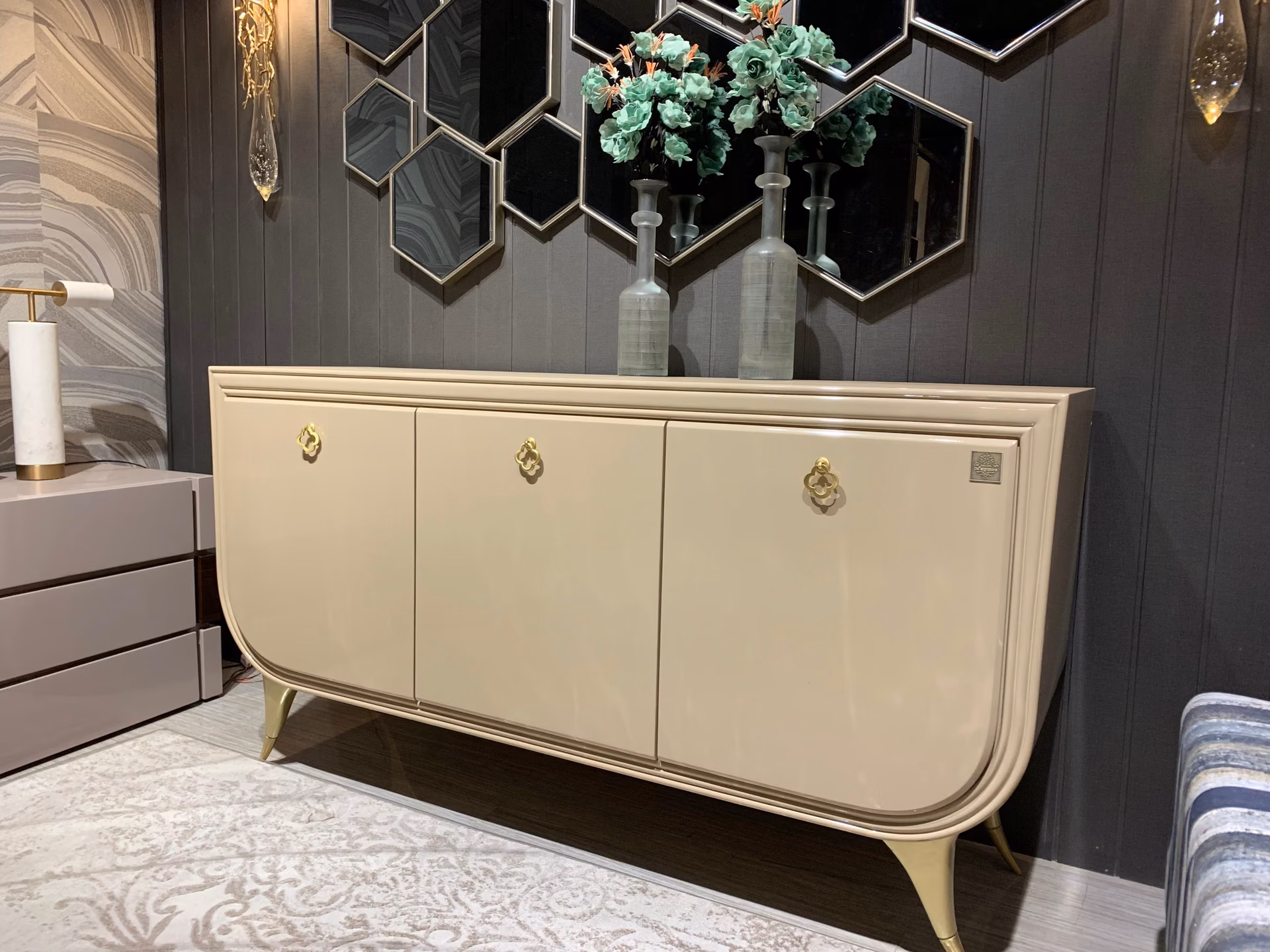 Champagne lacquer sideboard with curved ends, three doors, gold quatrefoil ring handles and splayed brass legs against a dark hexagon-mirror wall.