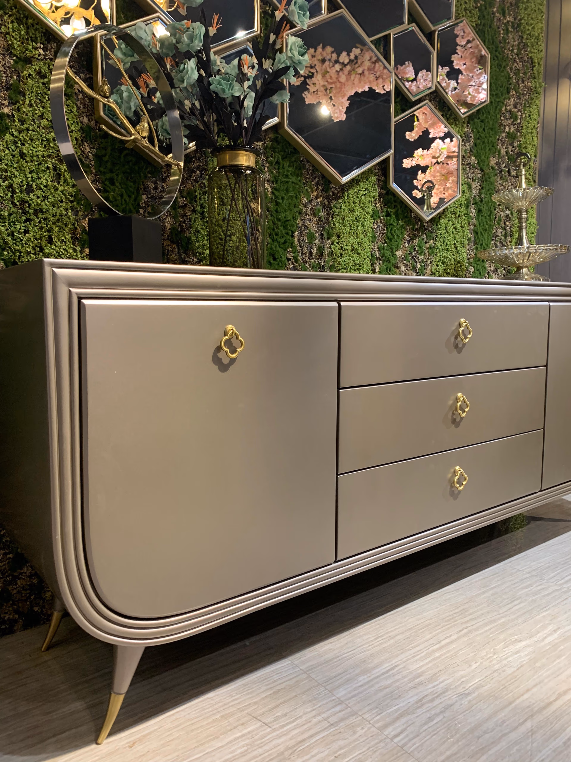 Angled view of the champagne lacquer sideboard showing a door and a bank of drawers with gold clover handles, set against a green moss wall with hexagon mirrors.