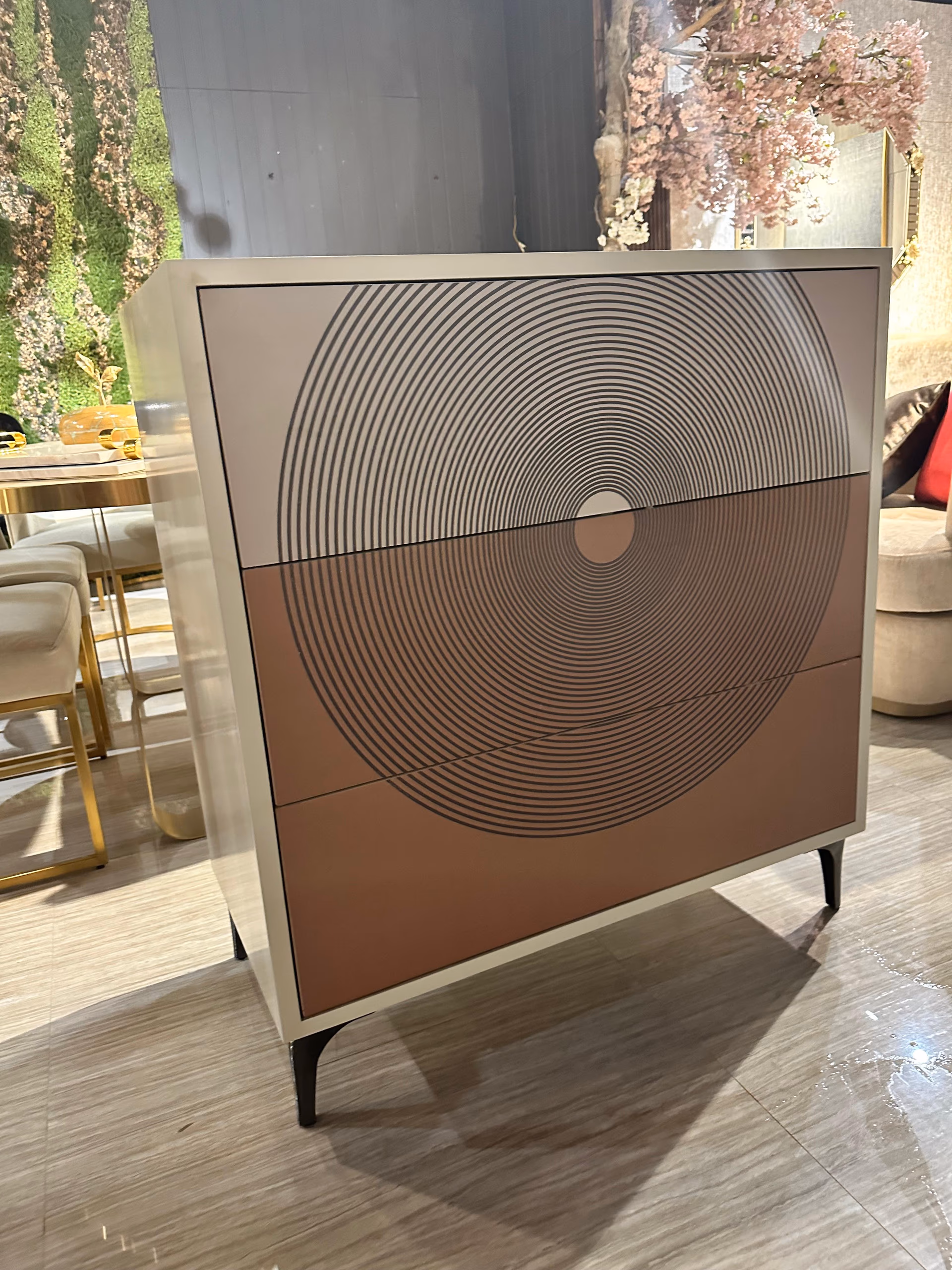 Tall cream crockery cabinet with a concentric-circle optical print across three drawer fronts on slim black legs.