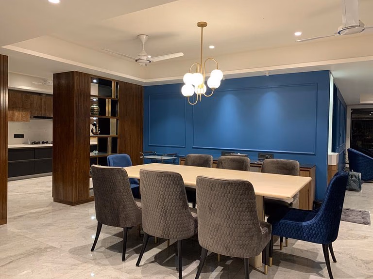 Eight-seat dining room with a cream stone-top table on a brass-trimmed base, grey and navy honeycomb-quilted chairs, a blue panelled feature wall and a fluted wood display niche.