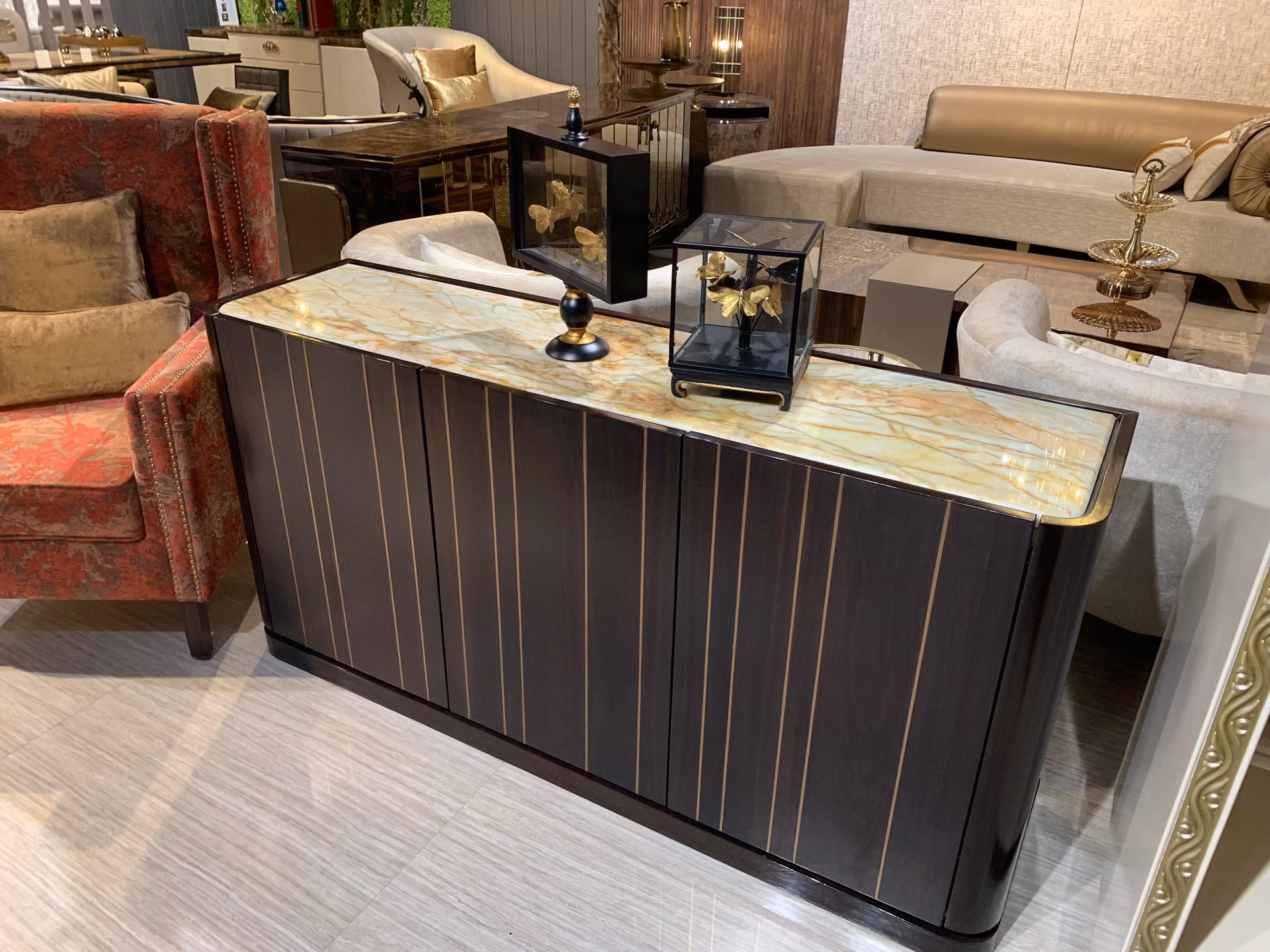 Corner view of the dark ebony fluted sideboard showing the onyx top overhang and brass-lined doors, beside a rust velvet chair in a showroom.