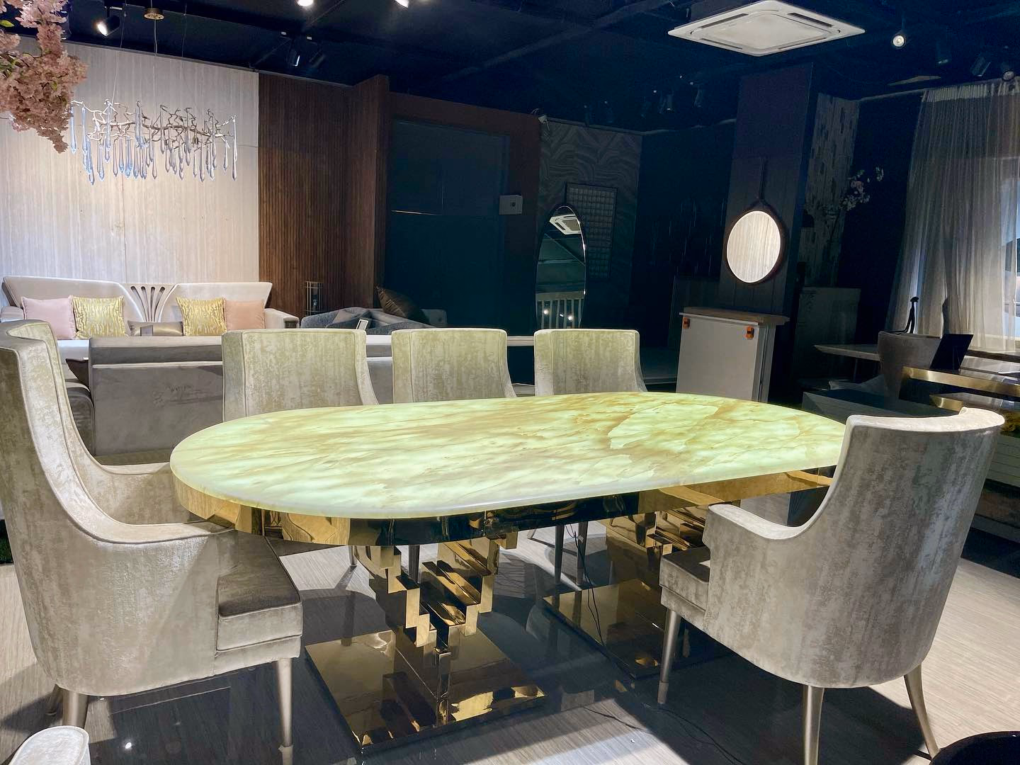Oval green onyx dining table on a sculpted stepped gold base, surrounded by taupe velvet armchairs on chrome legs in a lit showroom setting.