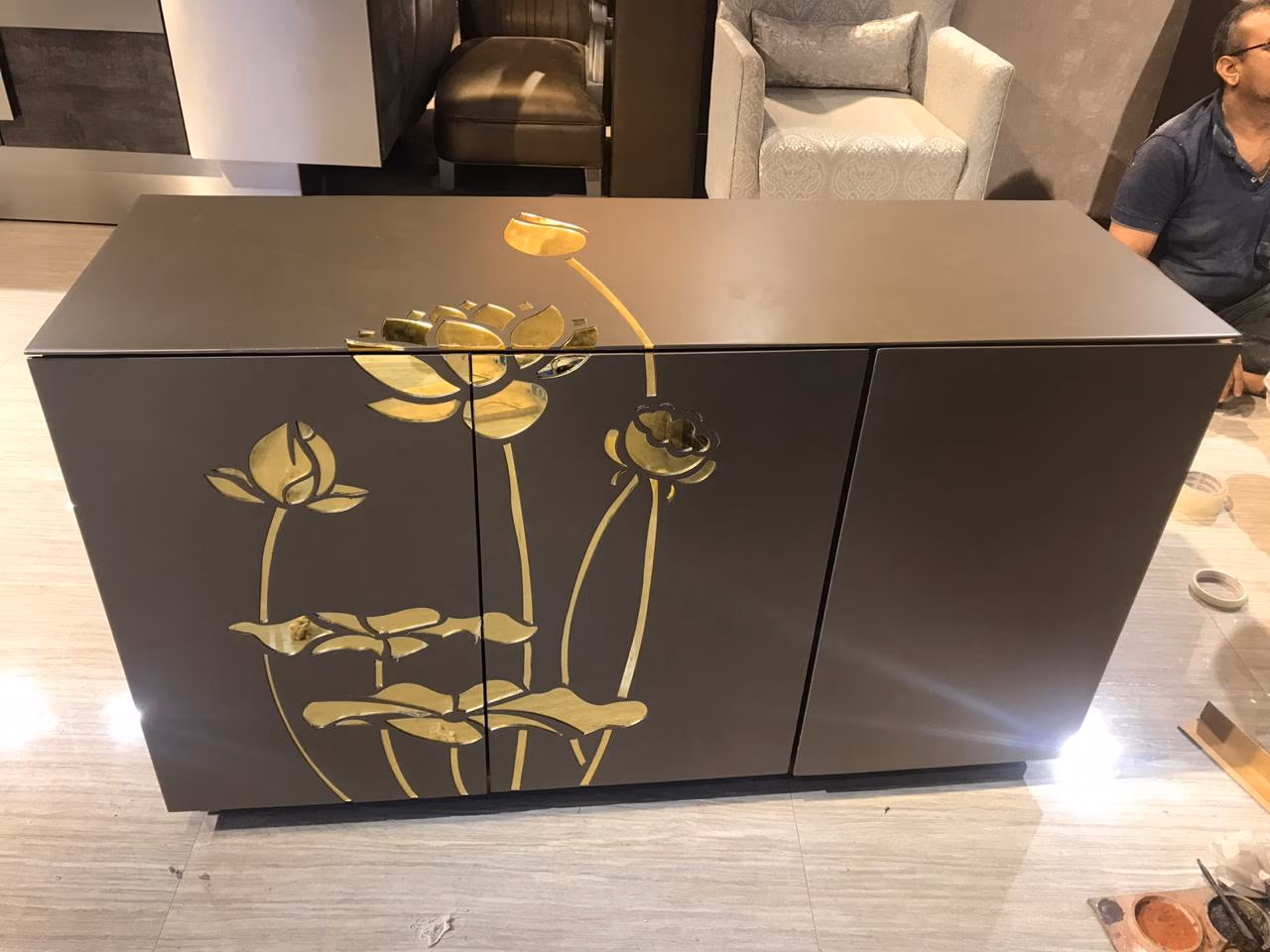 Low three-door sideboard in matte bronze-grey with a large hand-set brass lotus inlay spreading across the doors, standing on a workshop floor.