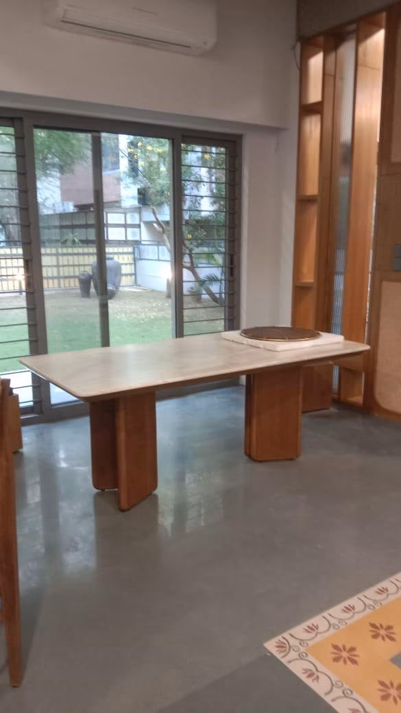 The marble-top dining table seen straight on in a bright room, its cream stone slab resting evenly on two curved solid-teak slab legs by a garden window.