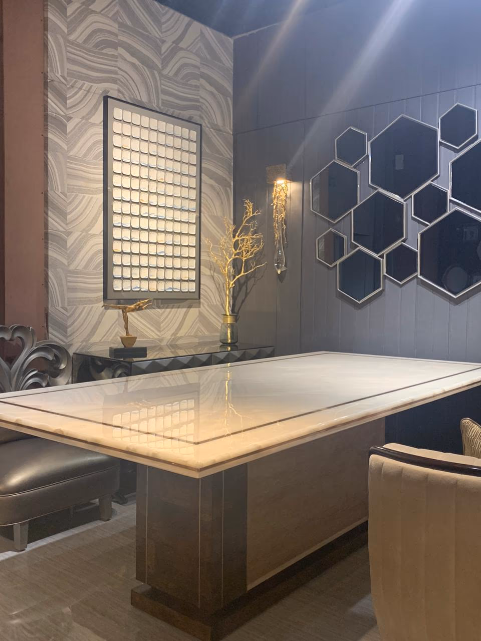 Wider view of the marble pedestal dining table in a styled room with hexagon mirror art, a beaded panel and a dark faceted console behind it.
