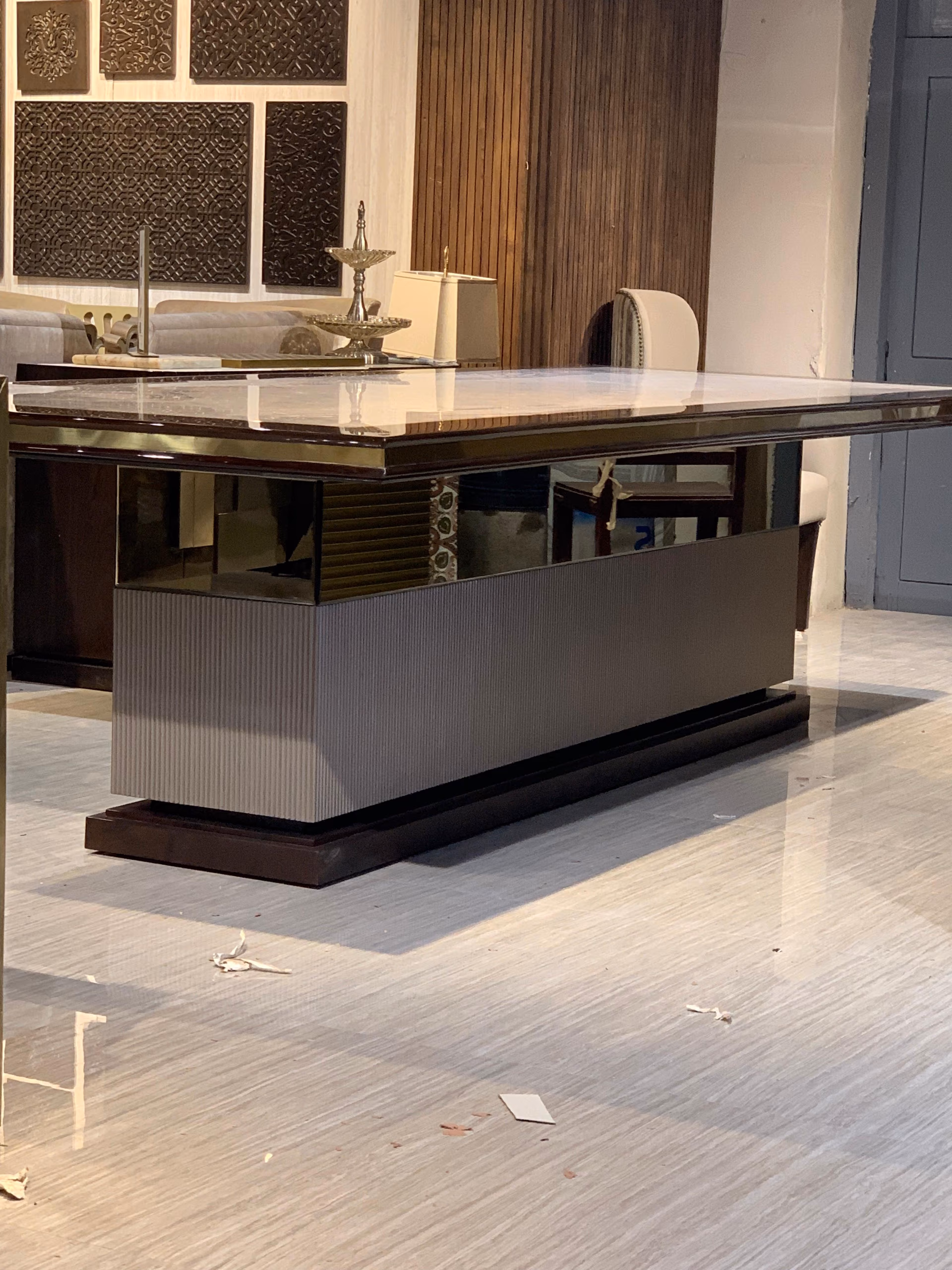 Large table with a beige marble top and glossy dark wood border, raised on a grey fluted pedestal over a dark plinth, in a showroom with carved wall panels.