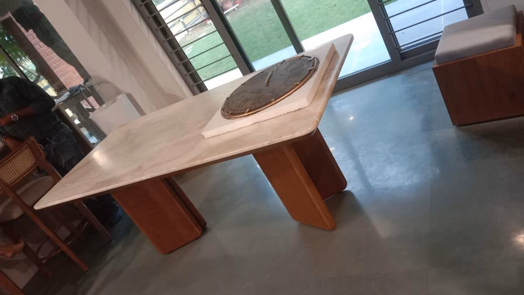 Rectangular dining table with a thick cream marble top and two curved solid-teak slab legs, holding a bronze and stone lazy-susan tray, on a concrete floor.